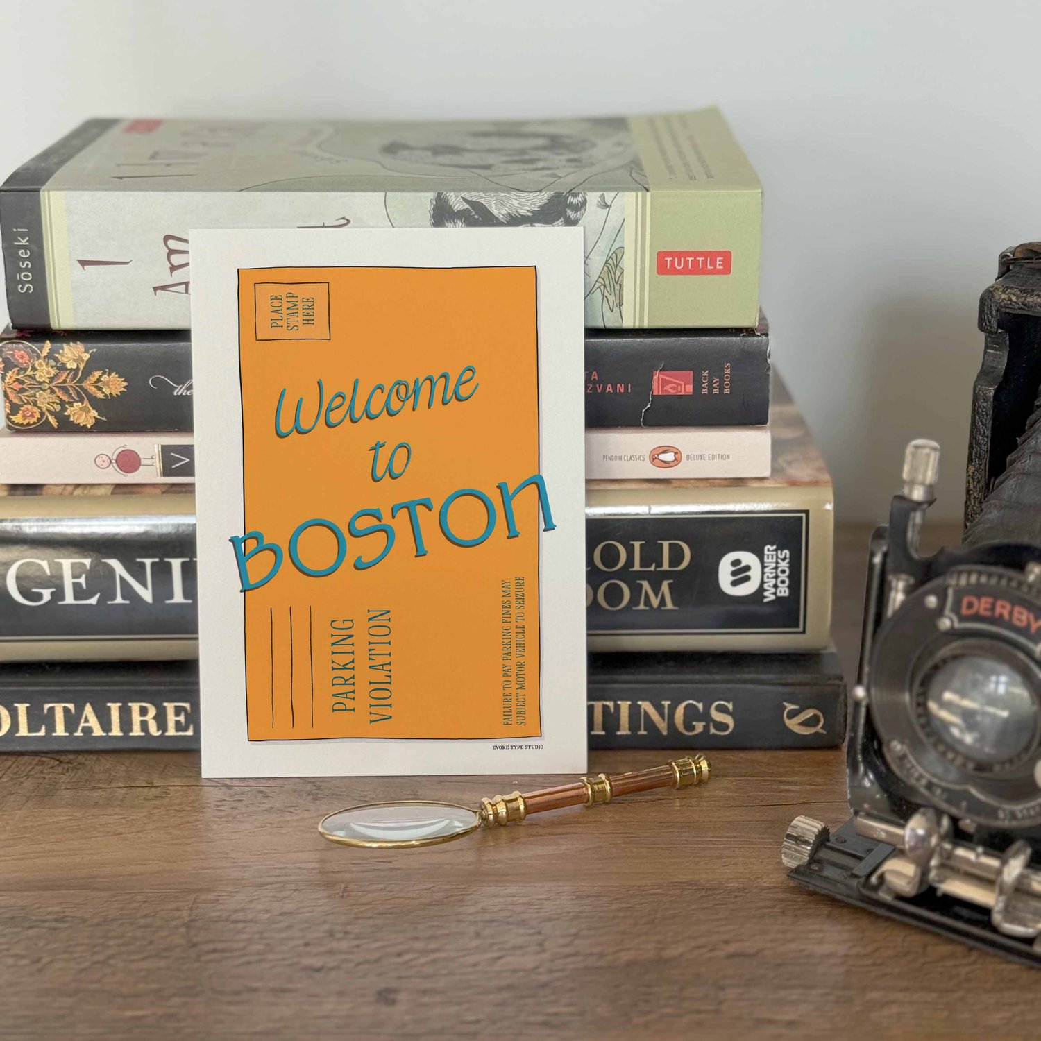 A notecard placed in front of a stack of books with a vintage camera and a small magnifying glass placed nearby. The artwork on the notecard is of an orange parking ticket envelope with the words "Welcome to Boston" drawn in unique lettering styles.