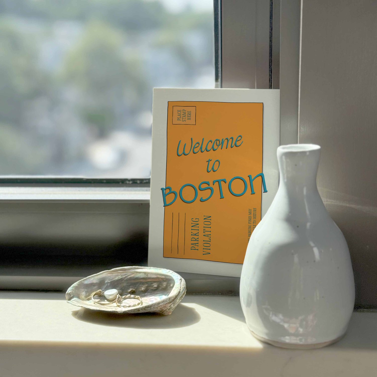A notecard sitting on a windowsill surrounding by a white vase and a pretty shell with jewelry placed inside.