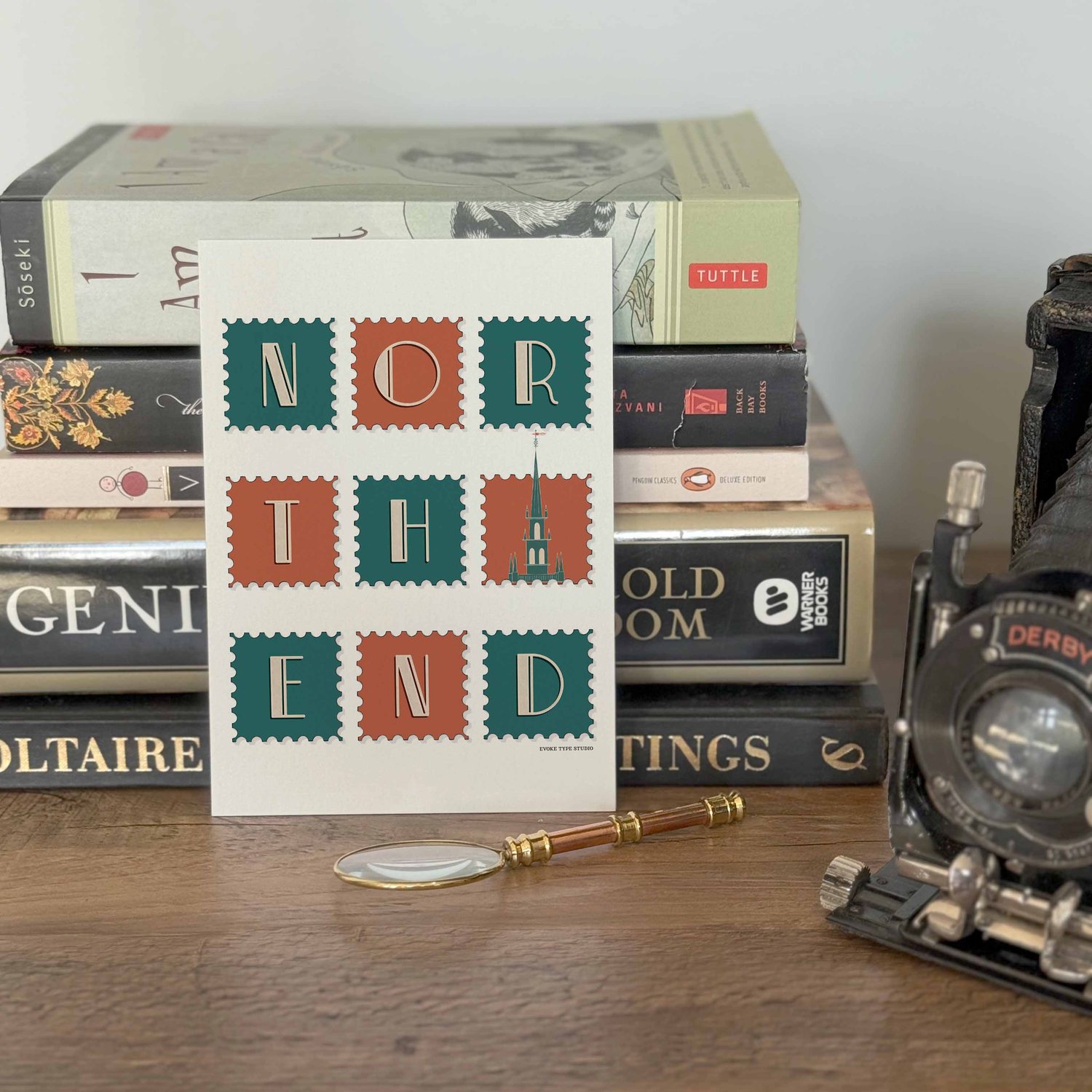 A notecard placed in front of a stack of books with a vintage camera and a small magnifying glass placed nearby. The artwork on the notecard is of postage stamps featuring the North End neighborhood in Boston, Massachusetts drawn in a unique lettering sty