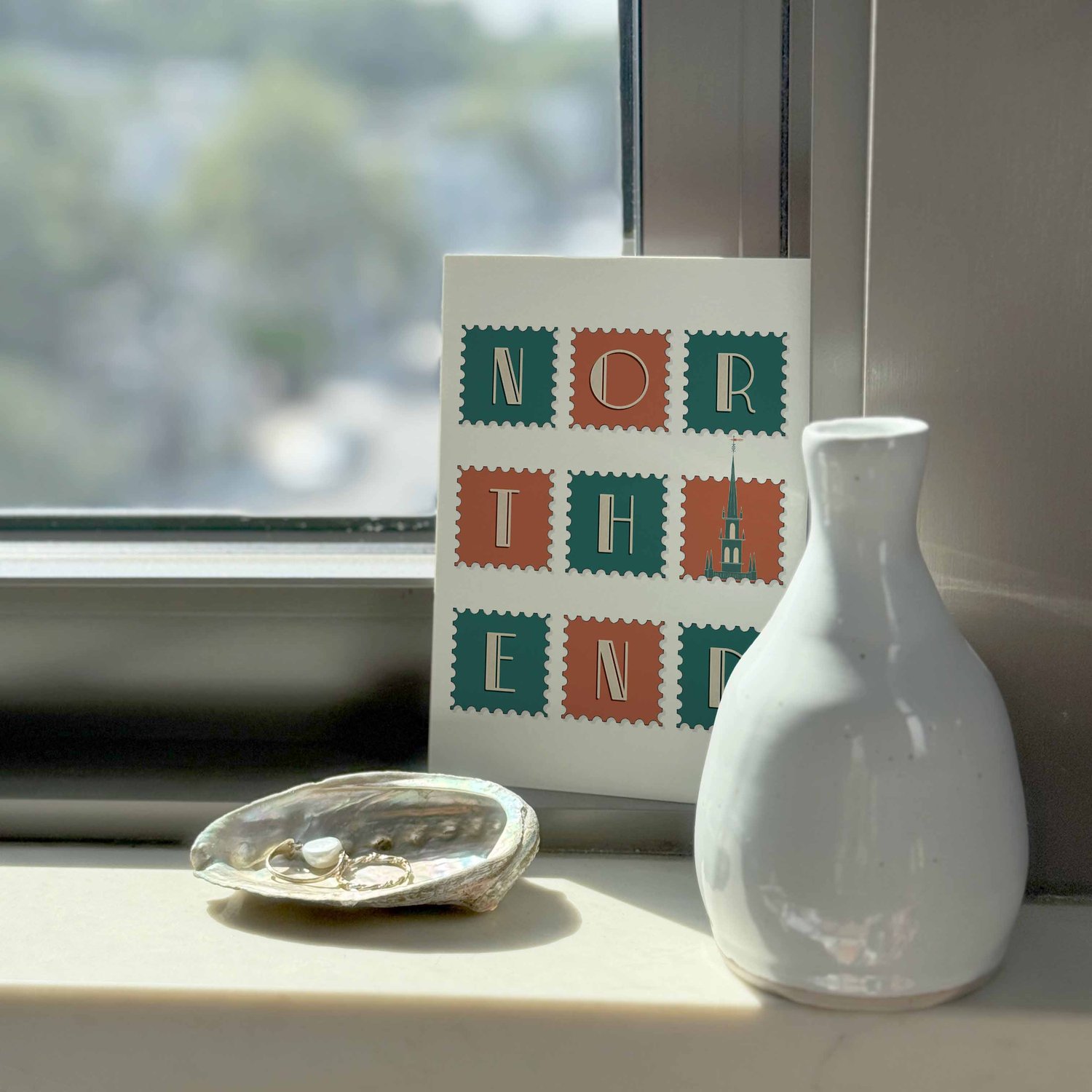 A notecard sitting on a windowsill surrounding by a white vase and a pretty shell with jewelry placed inside.