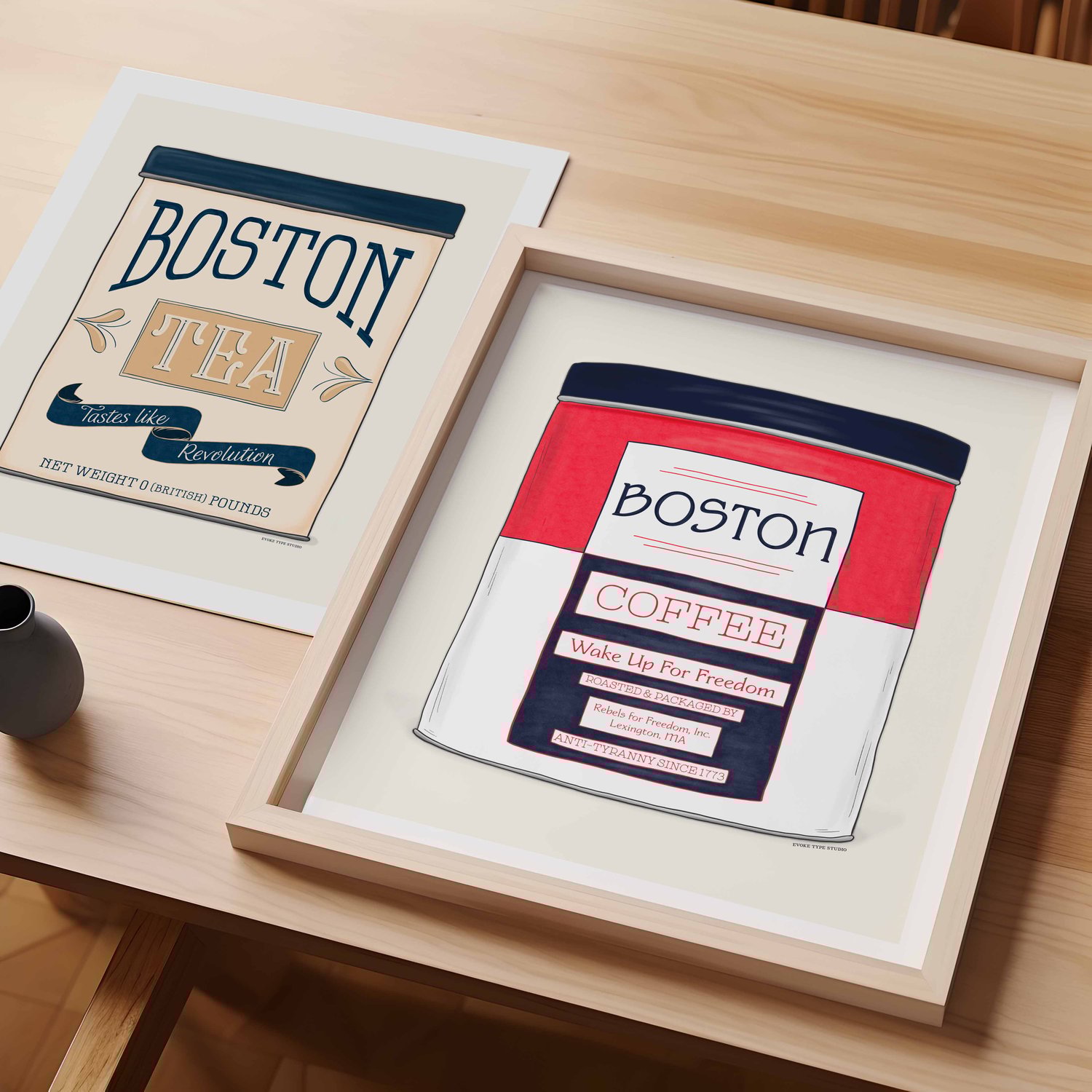 A table with an unframed artwork of a fake vintage Boston Tea Tin and a framed artwork of fake vintage Boston Coffee Tin.