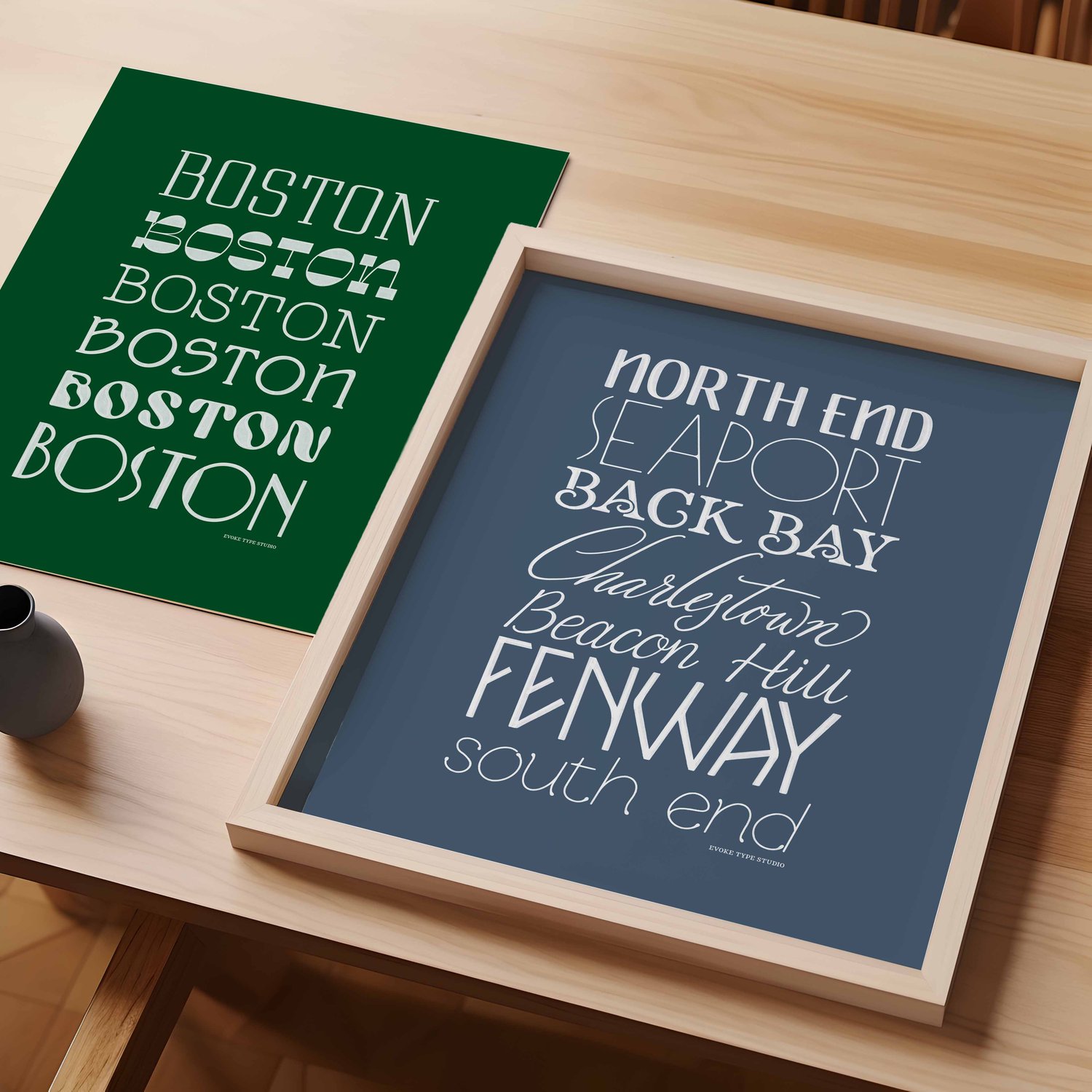 A table with an unframed artwork of the word "Boston" drawn in various lettering styles and a framed artwork of several Boston neighborhoods drawn in various lettering styles.