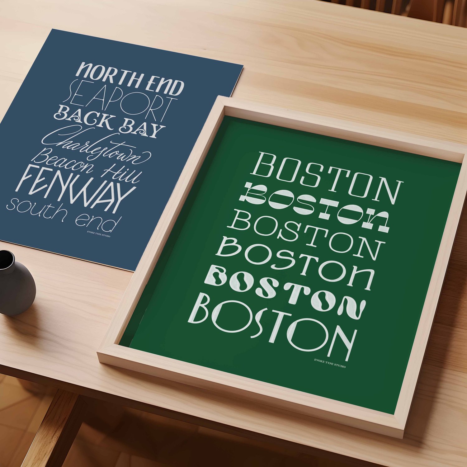 A table with an unframed artwork of several Boston neighborhoods drawn in various lettering styles and a framed artwork of the word "Boston" drawn in various lettering styles