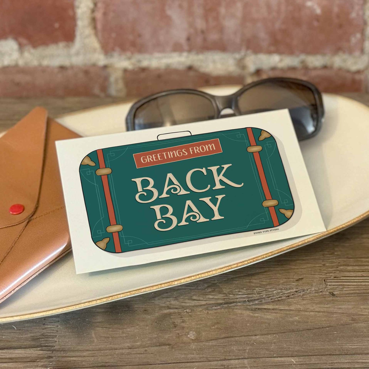 A postcard with a vintage suitcase illustration placed on a bureau with a wallet and sunglasses placed nearby.