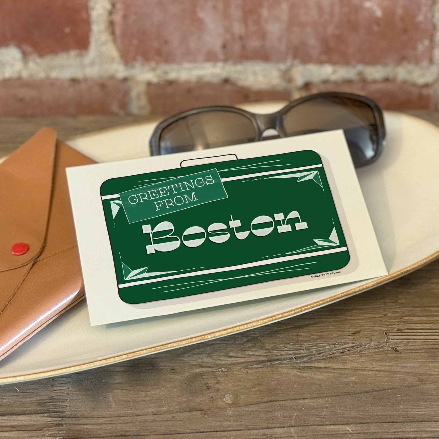 A postcard with a vintage suitcase illustration placed on a bureau with a wallet and sunglasses placed nearby.