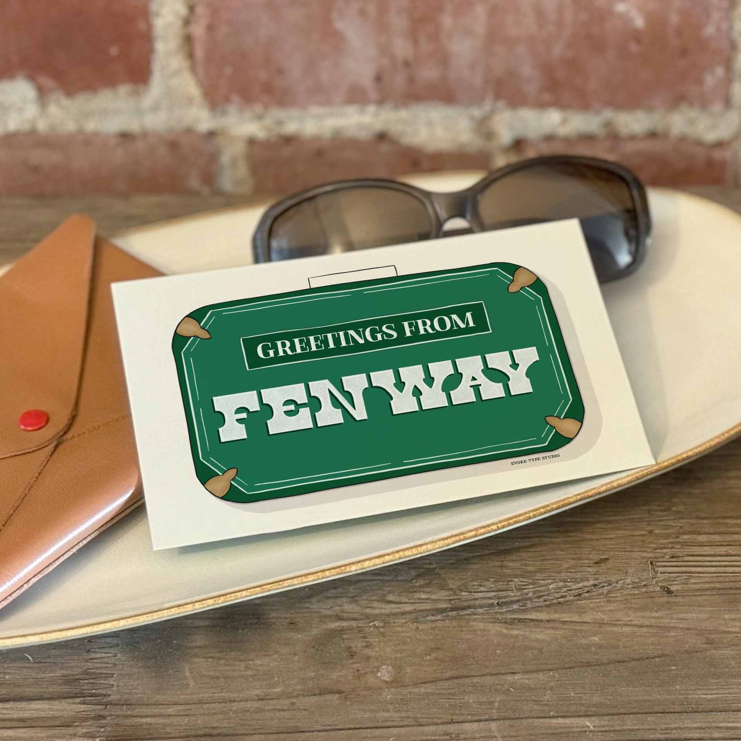 A postcard with a vintage suitcase illustration placed on a bureau with a wallet and sunglasses placed nearby.