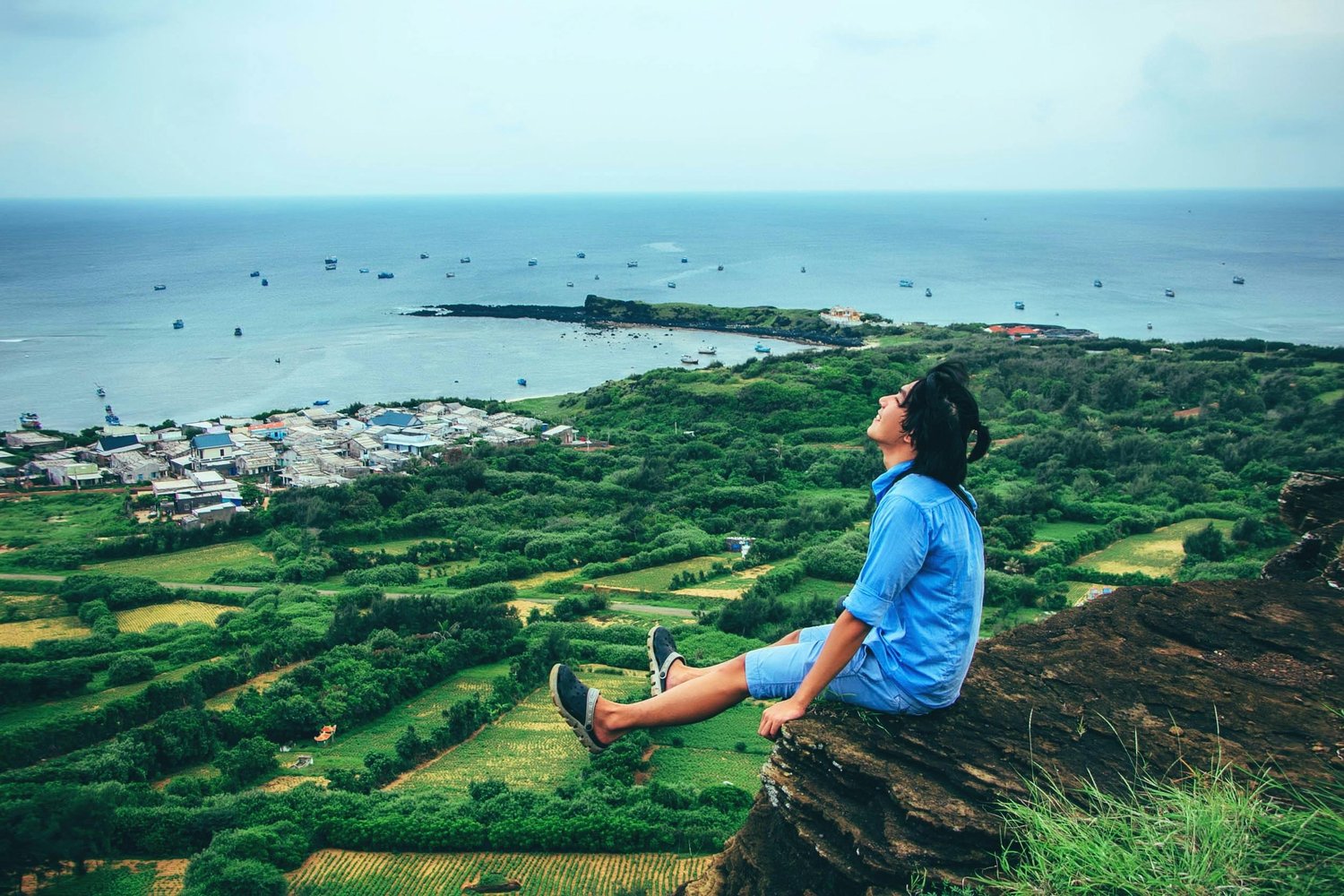 happy young man sitting on a mountain overlooking a green pasture leading to the vast ocean