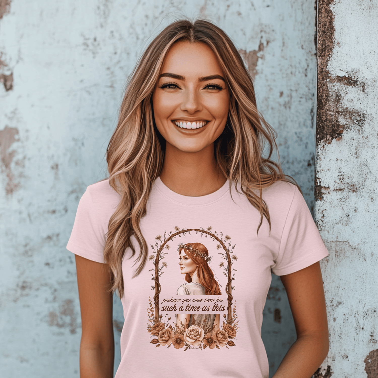 Smiling woman wearing a light pink T-shirt with a vintage-style graphic of a red-haired woman in a floral crown framed by roses and branches, featuring the inspirational quote 'Perhaps you were born for such a time as this,' standing against a rustic blue