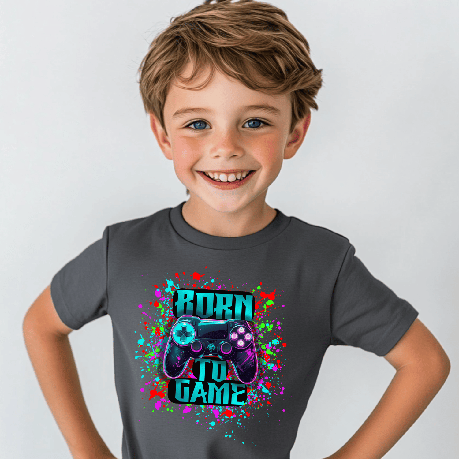 Smiling young boy wearing a dark gray T-shirt featuring a colorful splatter design with a neon video game controller and the bold text 'Born to Game,' standing against a light gray background.