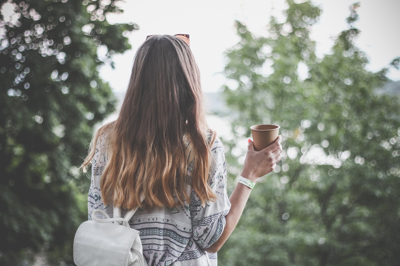 A childfree woman facing away, toward the trees, while holding a coffee in her right hand, up toward her shoulder.