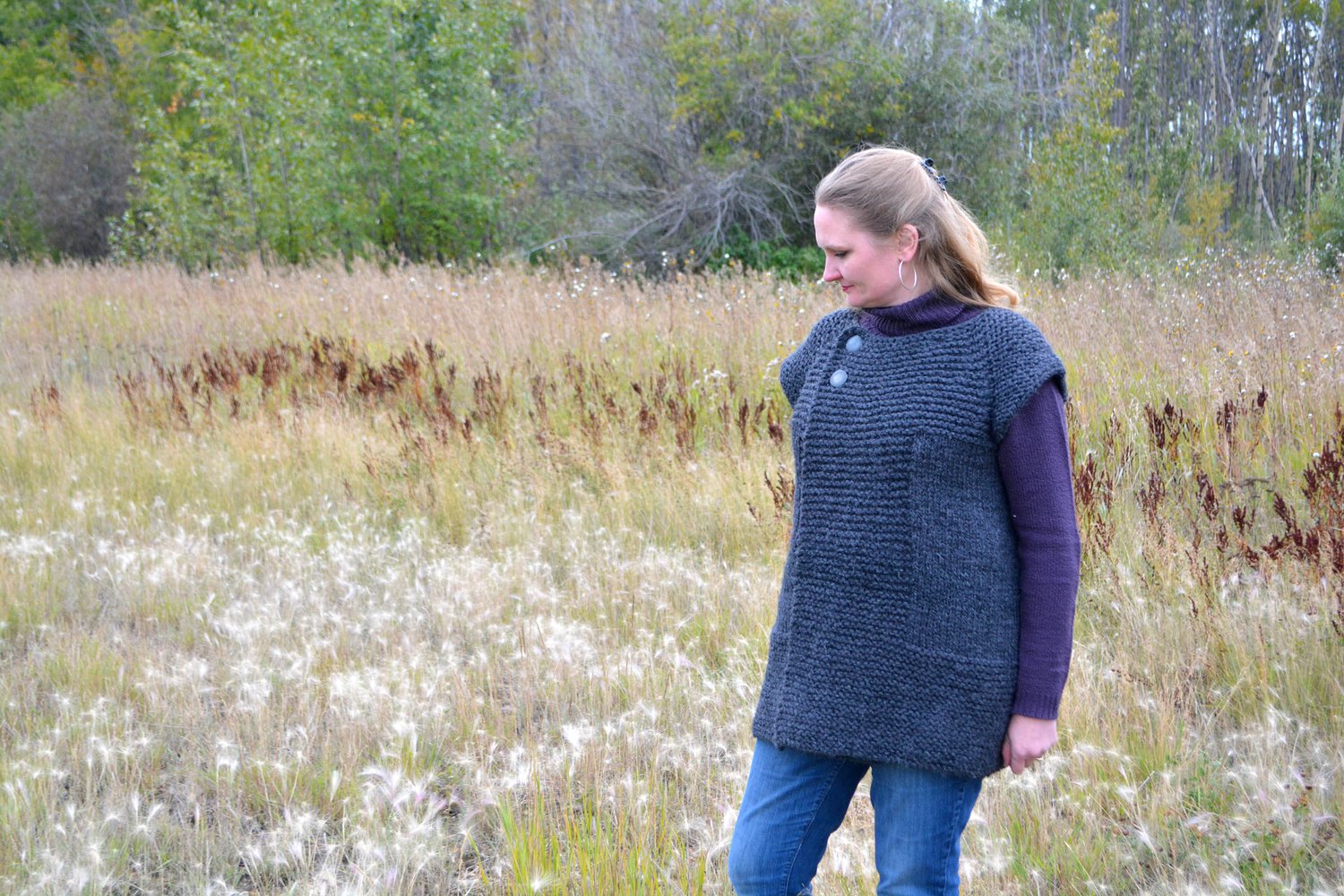 Talena wears a chunky-knit sleeveless coat in a field of prairie grasses.