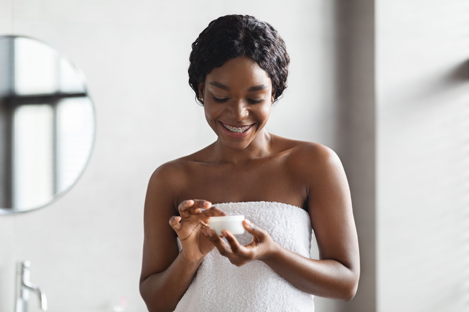 A woman wrapped in a white towel smiles while applying body lotion for dry skin from a small jar in a bright bathroom, with a round mirror in the background.