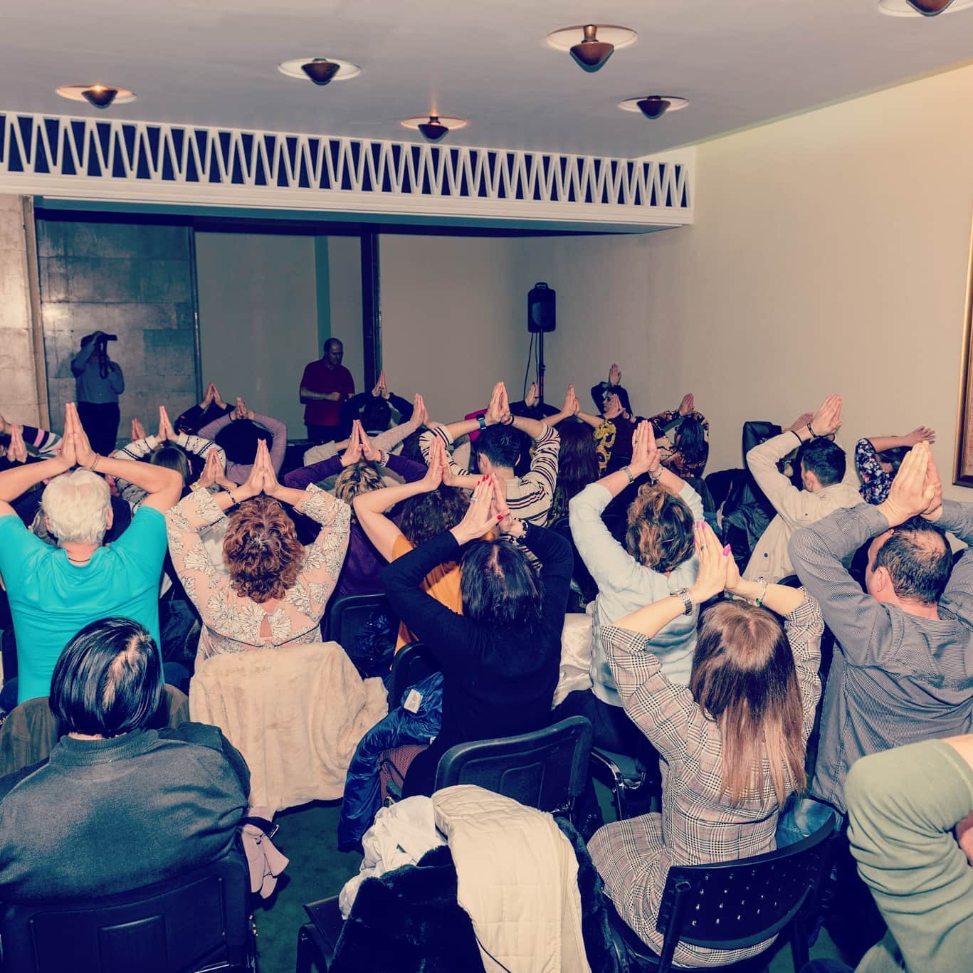 Grup de participanți într-o sală de conferință practicând o tehnică yoga ghidată de Eugen Mîrtz, în cadrul unui eveniment organizat de Alchimia Vieții – sesiune de reconectare spirituală și armonizare colectivă.