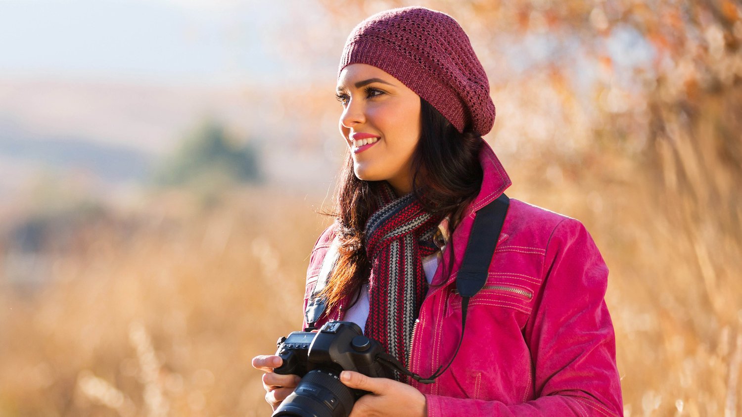 Young woman in a pink jacket and knitted hat holding a DSLR camera outdoors in golden light, ready to capture a landscape, featured in The Creative Guide Photography Blog.