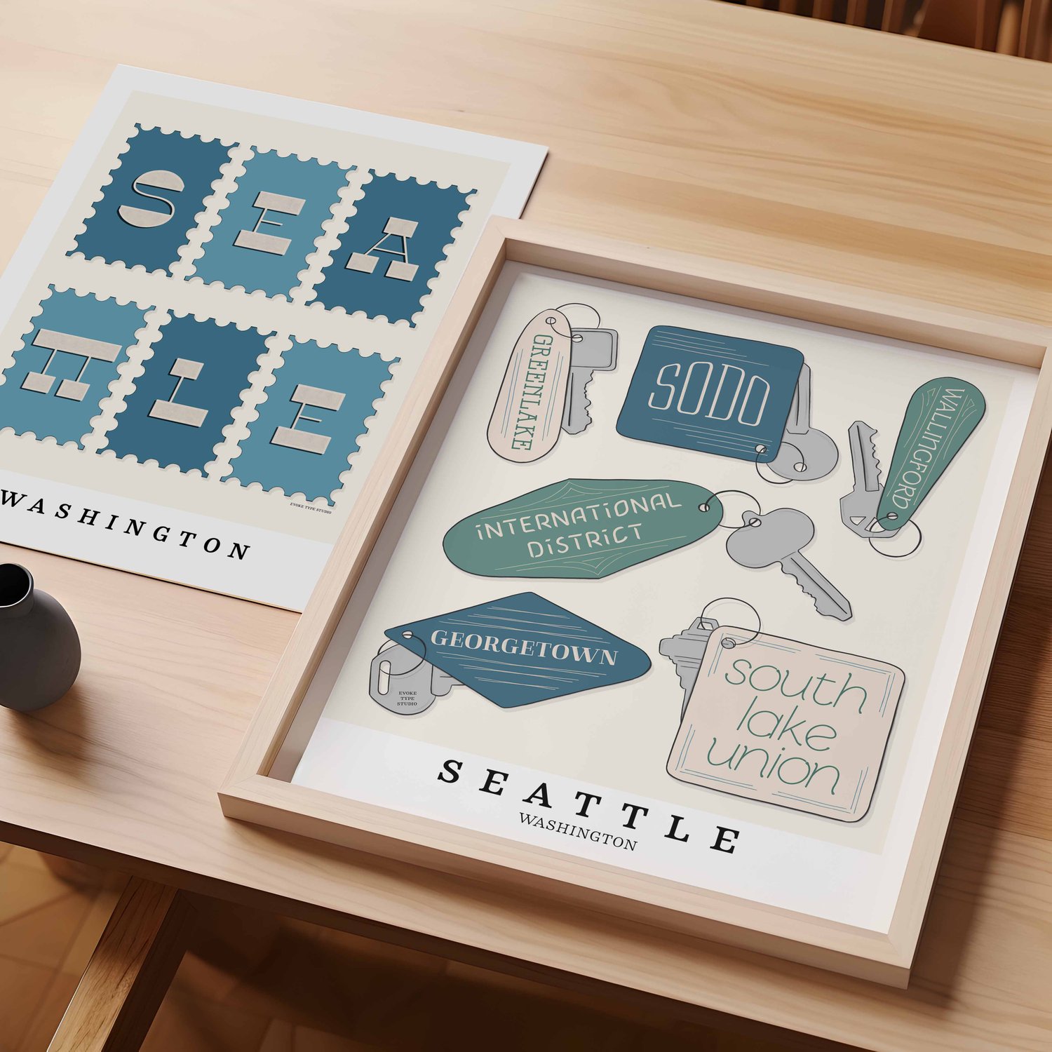 A table with an unframed artwork of Seattle postage stamp fob art and a framed artwork of Seattle key fob art