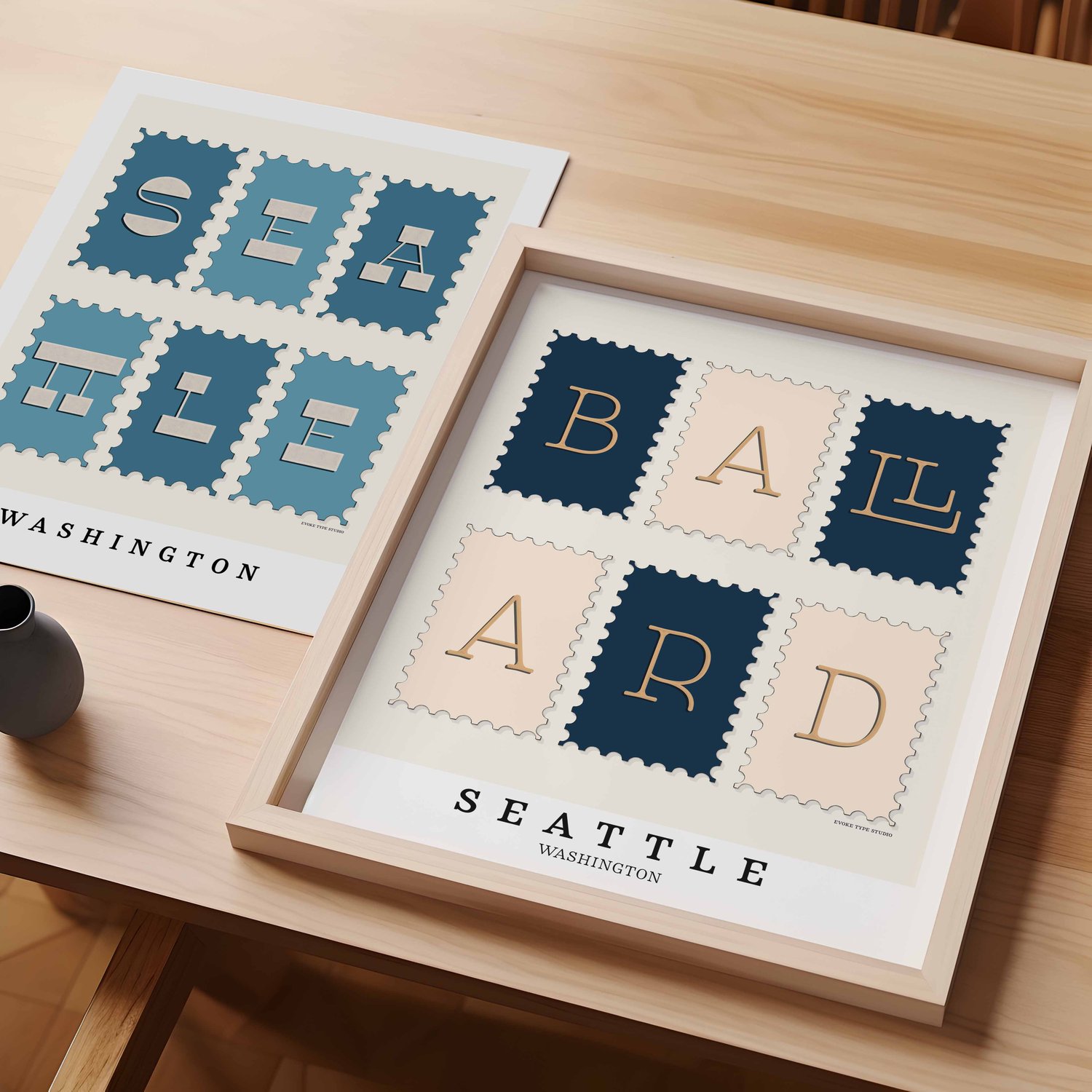 A table with an unframed artwork of Seattle postage stamp art and a framed artwork of Ballard postage stamp art