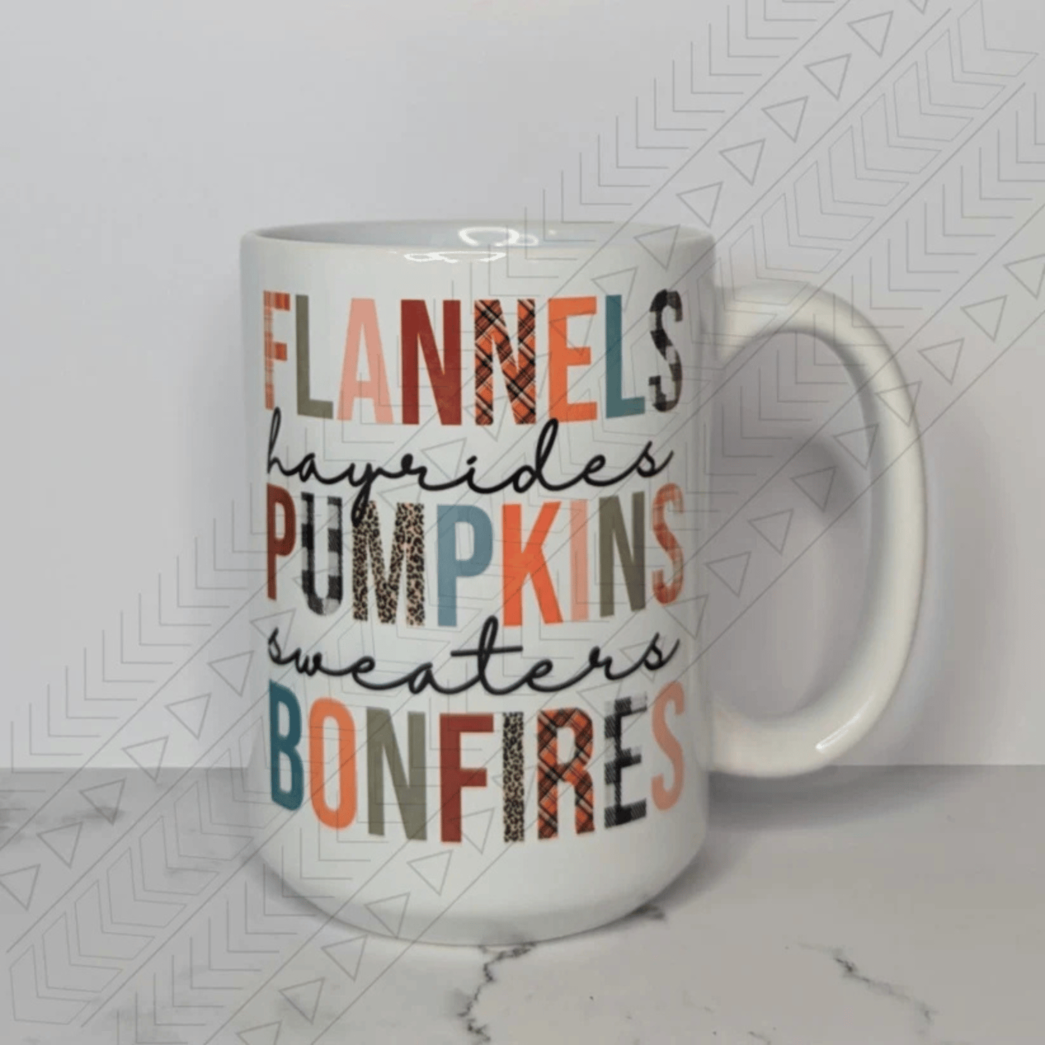 A white ceramic mug with the words "FLANNELS," "HAYRIDES," "PUMPKINS," "SWEATERS," and "BONFIRES" stacked vertically. The text is in a mix of different fonts and colors, including solid orange, green, and brown, as well as plaid and leopard print patterns