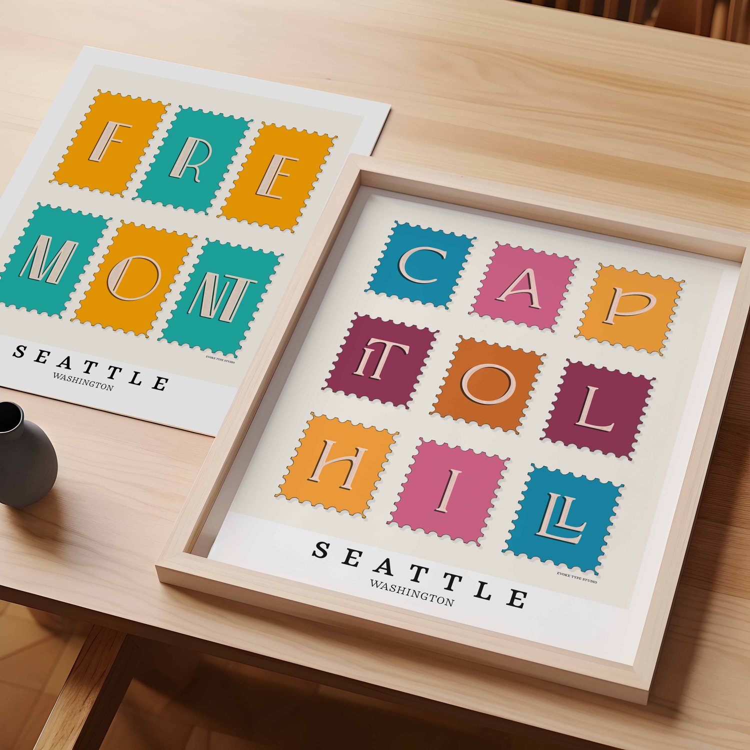 A table with an unframed artwork of Fremont postage stamp art and a framed artwork of Capitol Hill postage stamp art