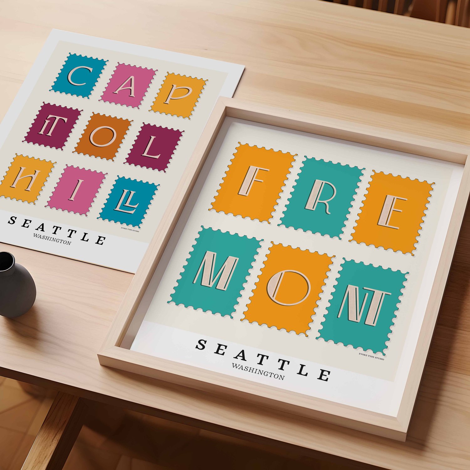 A table with an unframed artwork of Capitol Hill postage stamp art and a framed artwork of Fremont postage stamp art