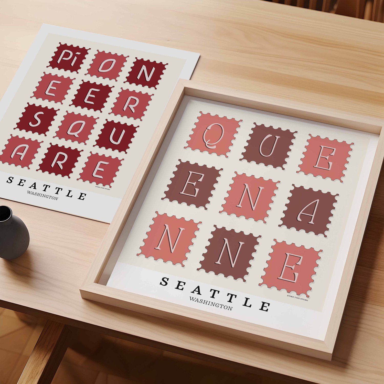 A table with an unframed artwork of Pioneer Square postage stamp art and a framed artwork of Queen Anne postage stamp art