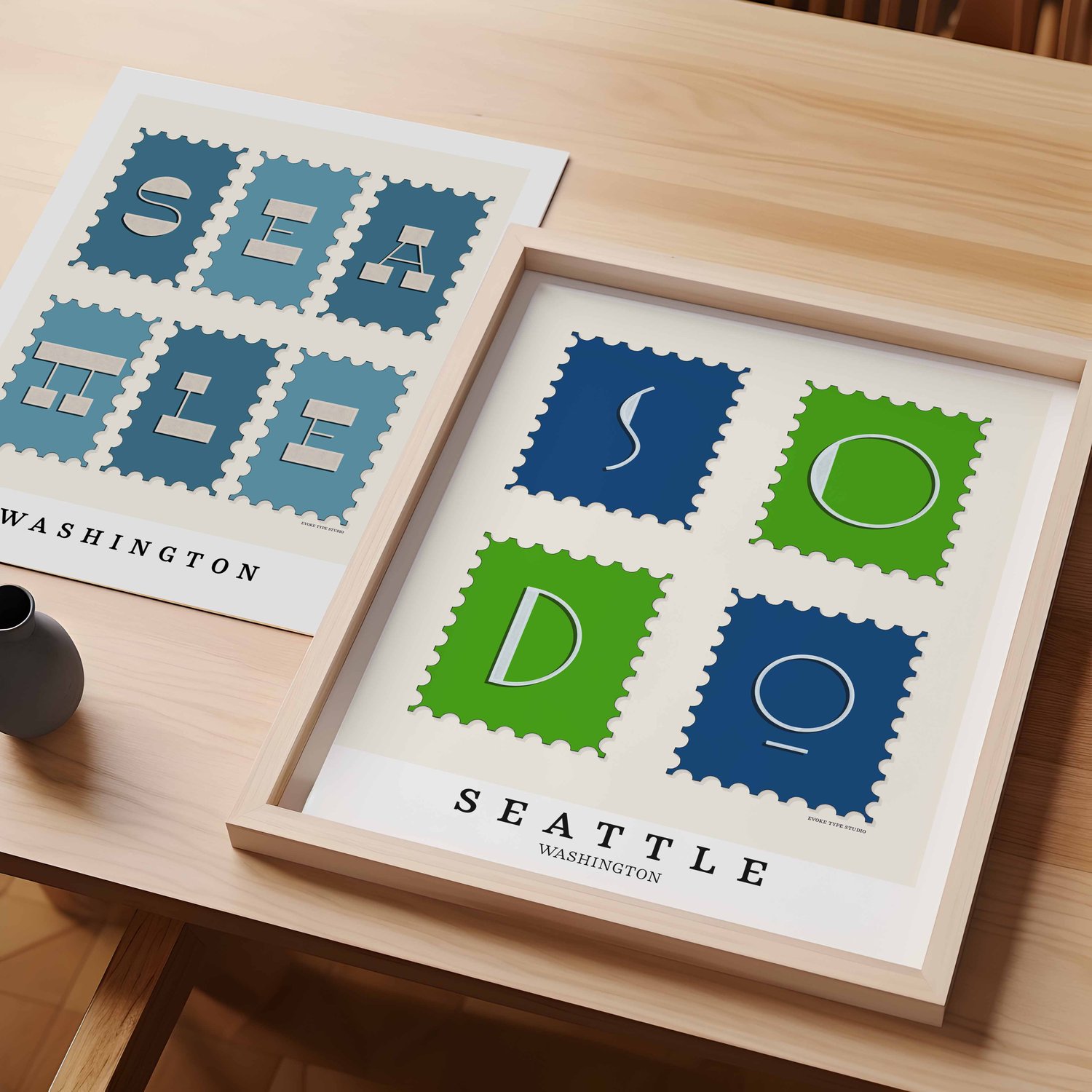 A table with an unframed artwork of Seattle postage stamp art and a framed artwork of SoDo postage stamp art