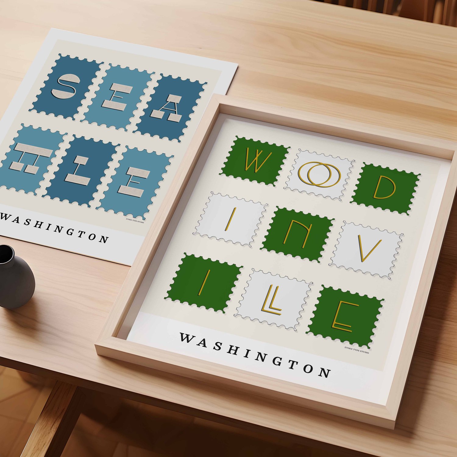 A table with an unframed artwork of Seattle postage stamp art and a framed artwork of Woodinville postage stamp art
