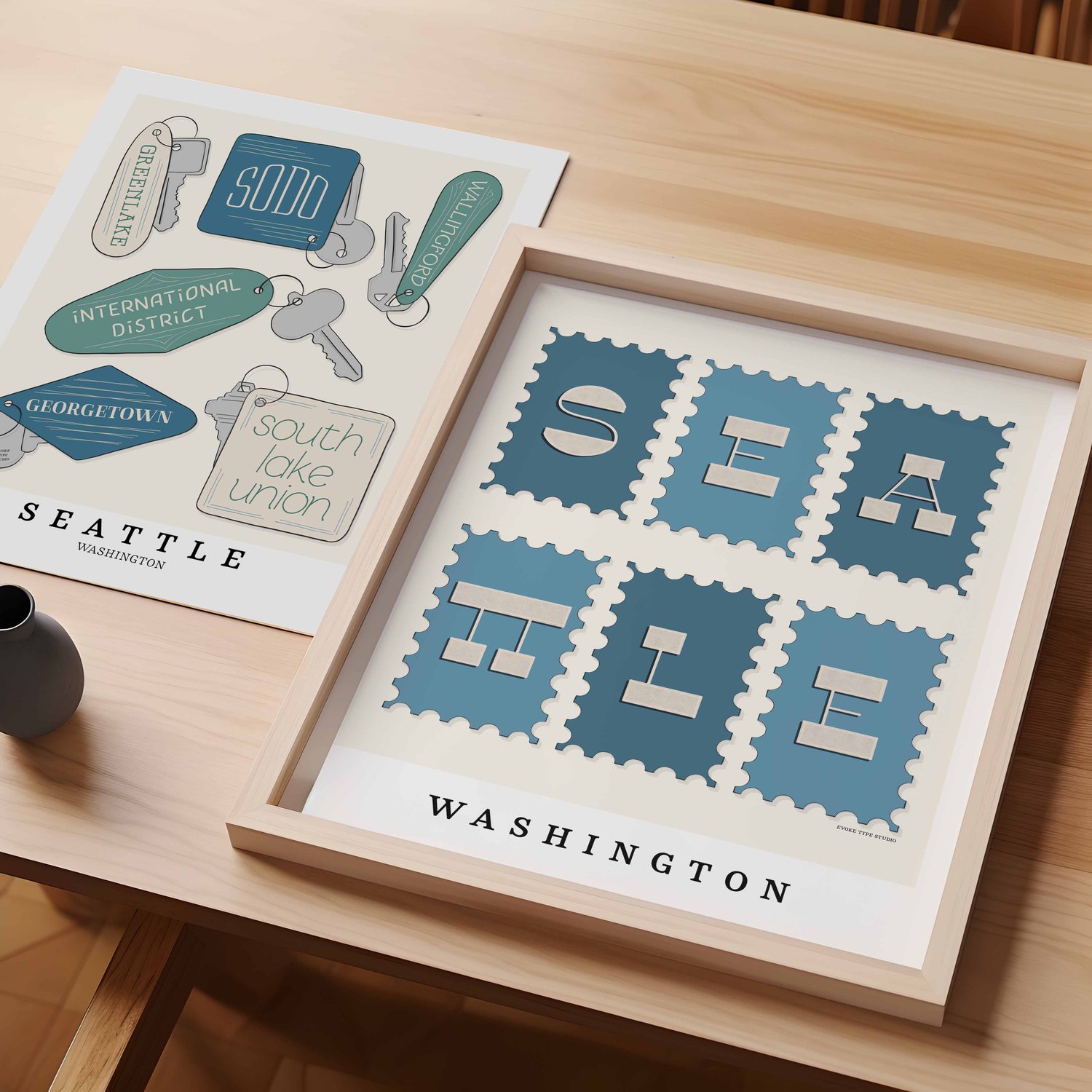A table with an unframed artwork of Seattle key fob art and a framed artwork of Seattle postage stamp art