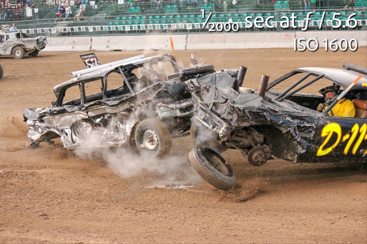 Demolition derby crash frozen mid-impact, with dust and debris flying as two heavily damaged cars collide, one losing a wheel, captured at 1/2000 sec, f/5.6, ISO 1600. Taken with a camera limited to a maximum ISO of 1600, using a lens zoomed fully in wher
