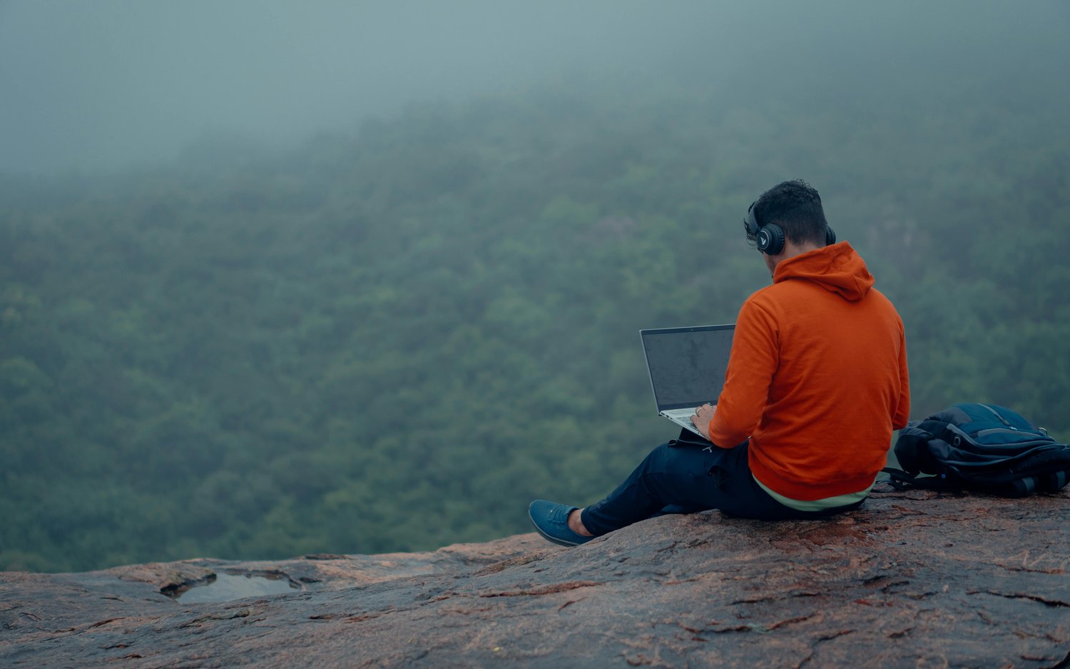 A man in an orange hoodie sits on a mountaintop overlooking the green forest while he does work on his laptop