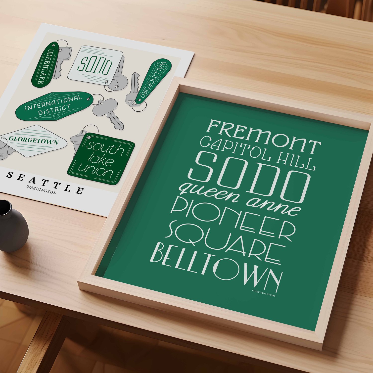 A table with an unframed artwork of Seattle, Washington key fobs drawn in various lettering styles and a framed artwork of several Seattle neighborhoods.