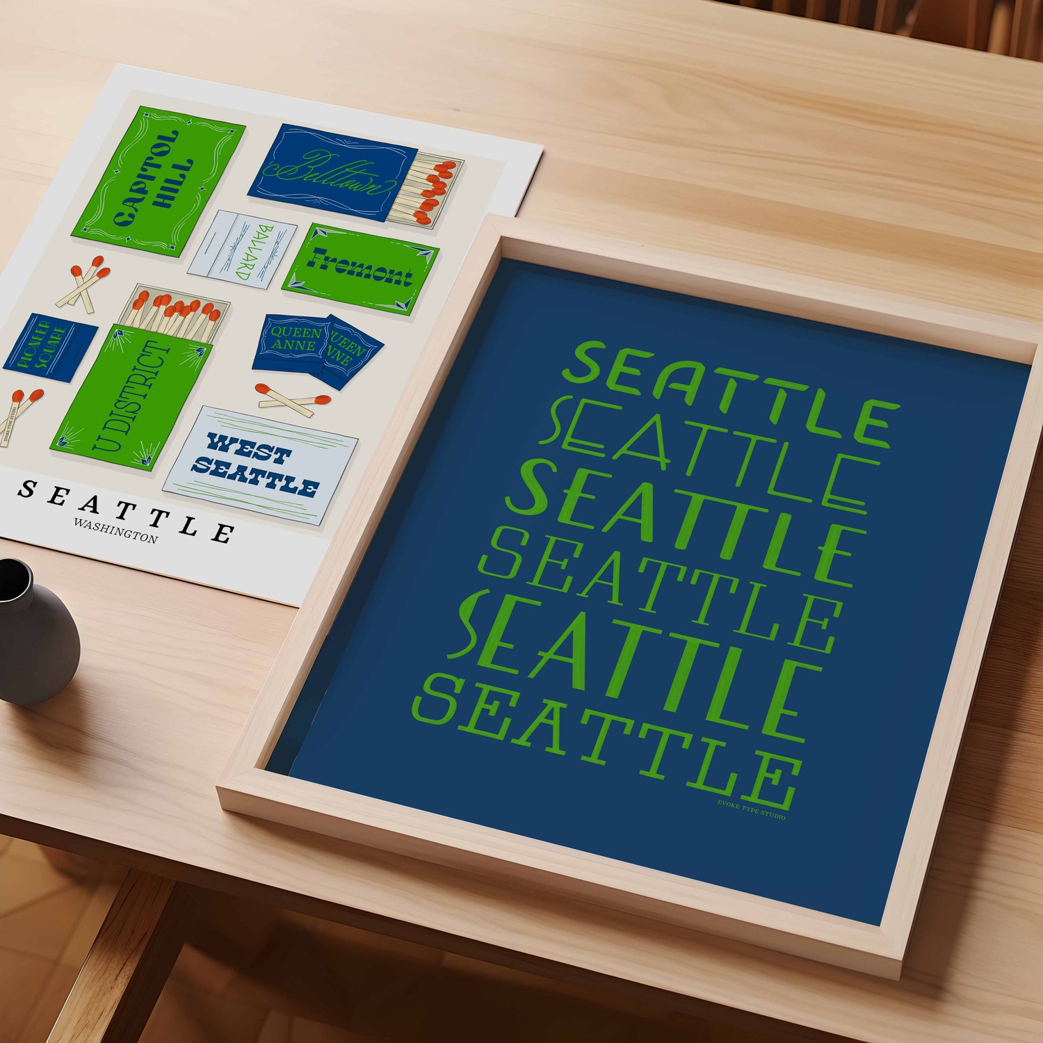 A table with an unframed artwork of Seattle, Washington neighborhoods drawn on matchboxes and a framed artwork of the word "Seattle" drawn in various lettering styles.