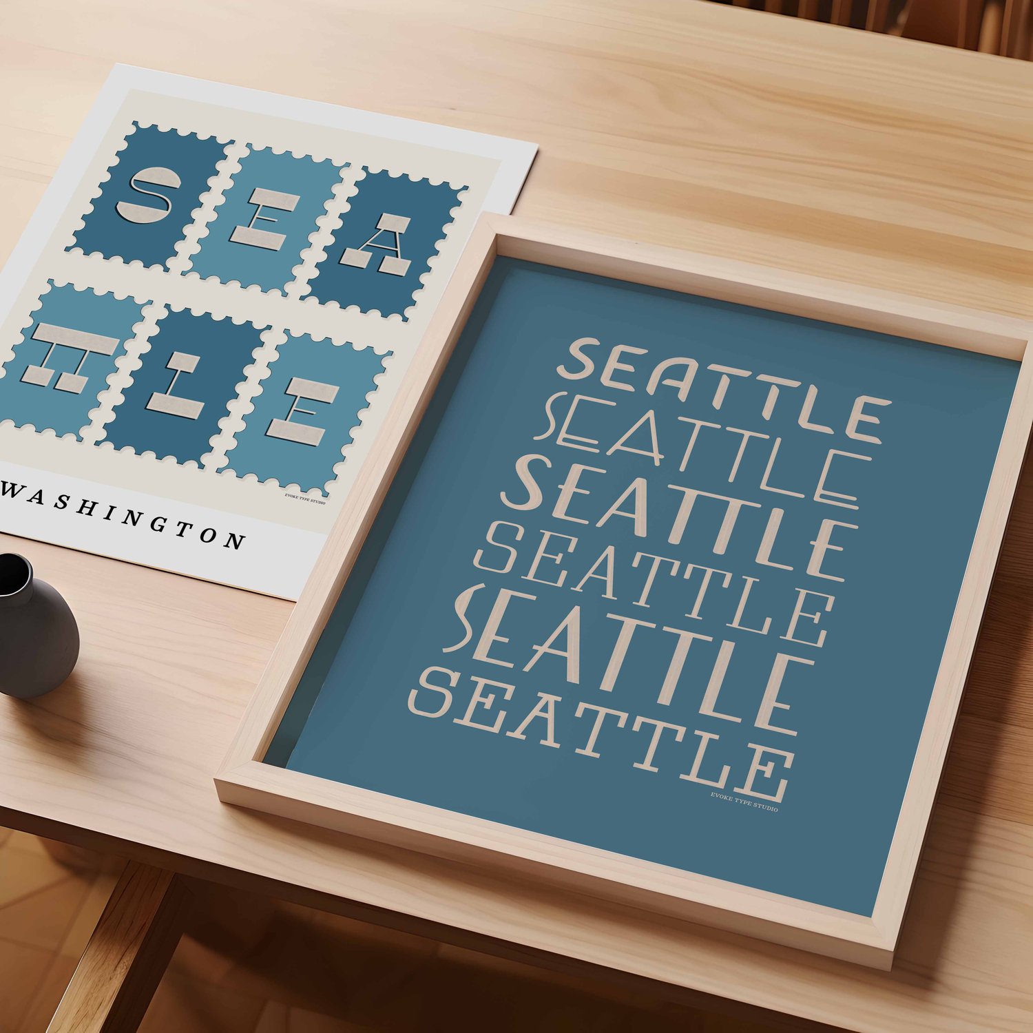 A table with an unframed artwork of the word "Seattle" drawn on postage stamps and a framed artwork of the word "Seattle" drawn in various lettering styles.