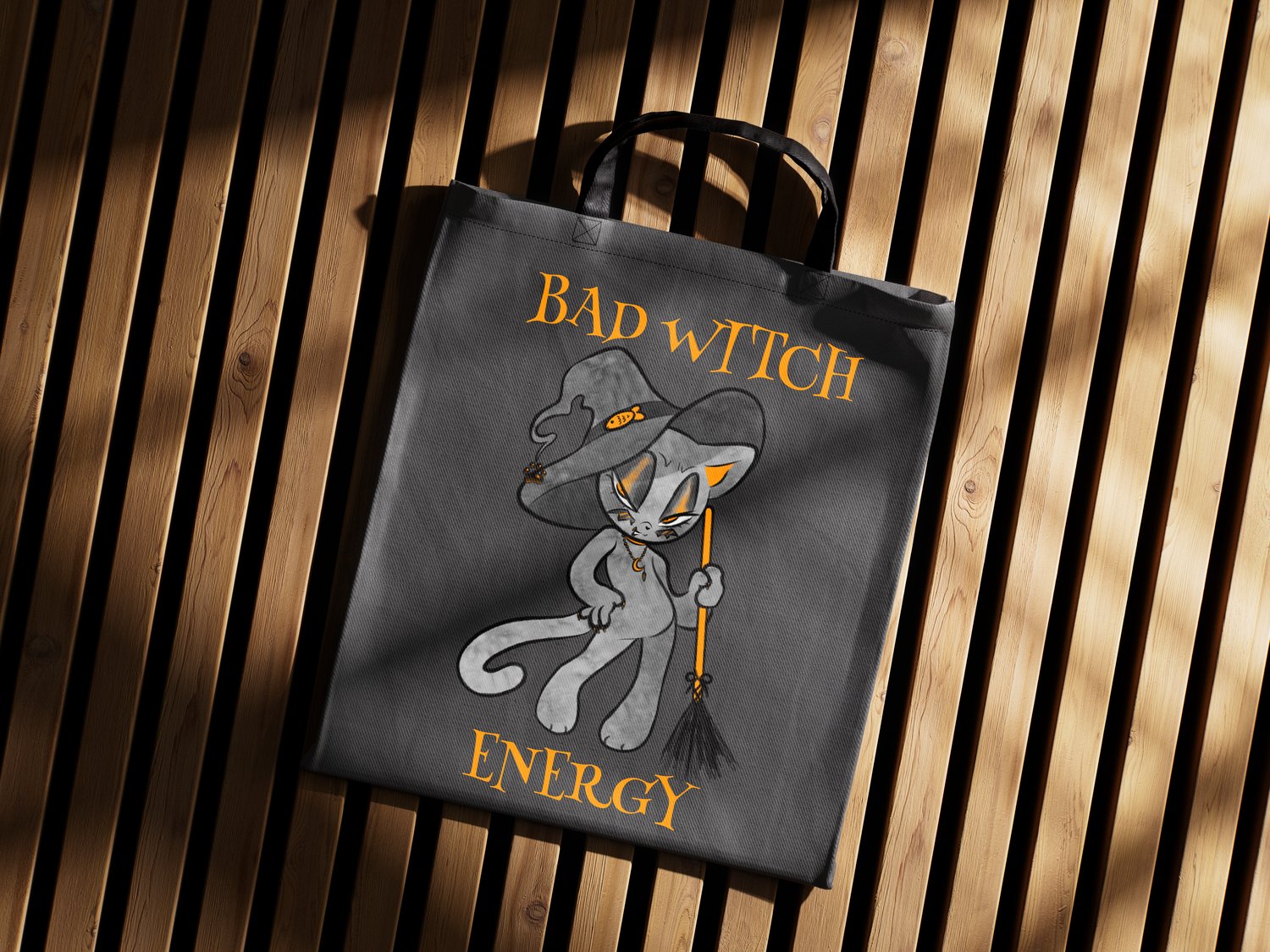 A dark grey tote bag with an illustration of a black and orange cat in a witch hat holding an orange broom. Orange text reads "Bad Witch Energy."