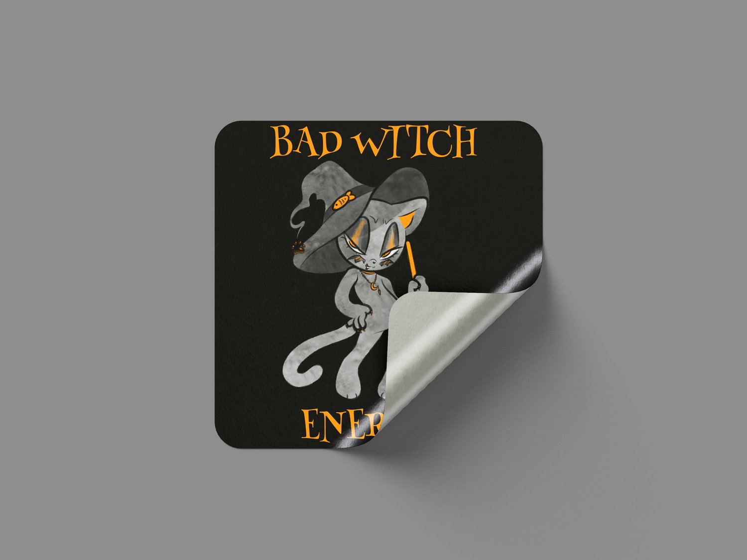 A black, square sticker on a grey background. The sticker is being peeled up and has an illustration of a black and orange cat in a witch hat holding an orange broom. Orange text reads "Bad Witch Energy."