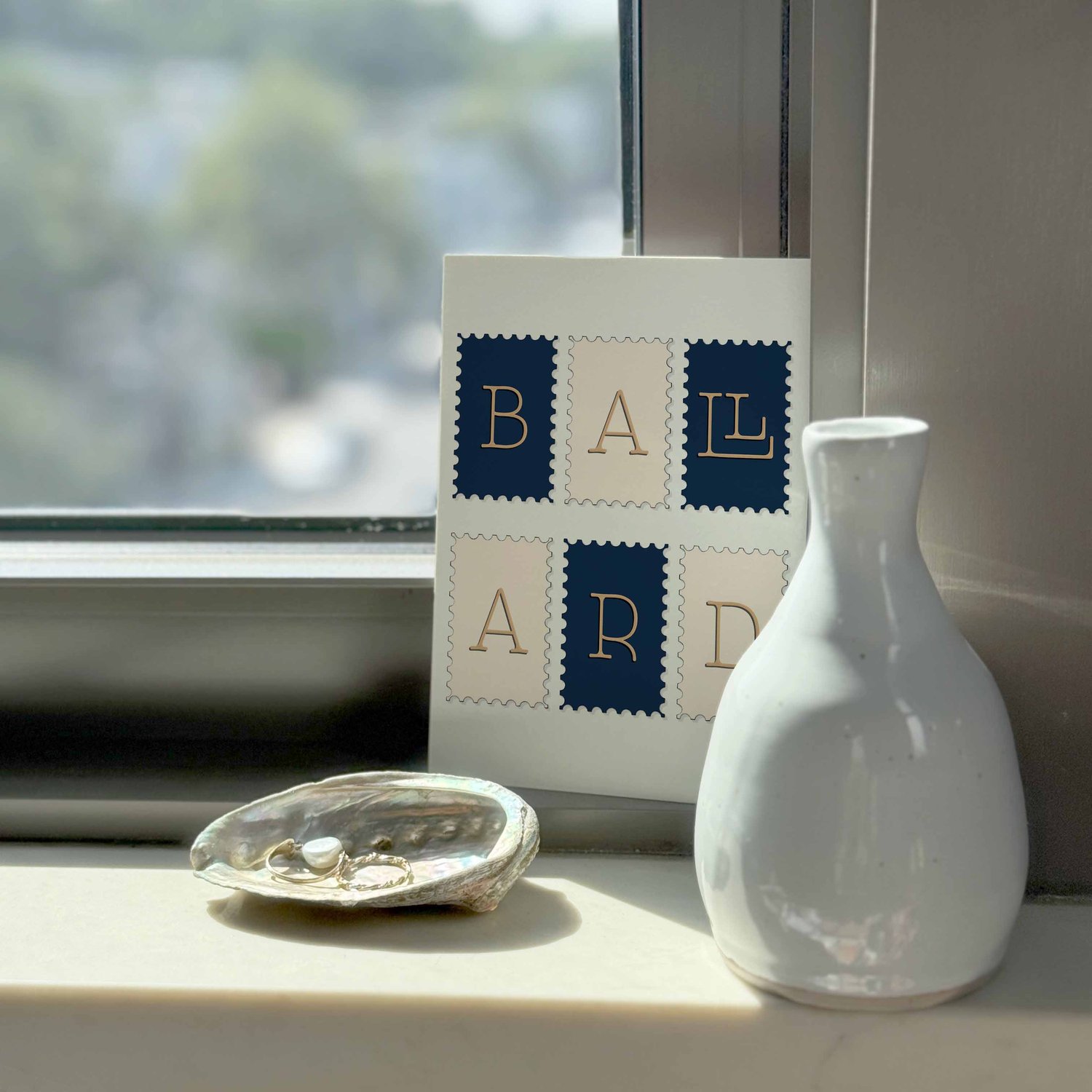 A notecard sitting on a windowsill surrounding by a white vase and a pretty shell with jewelry placed inside.