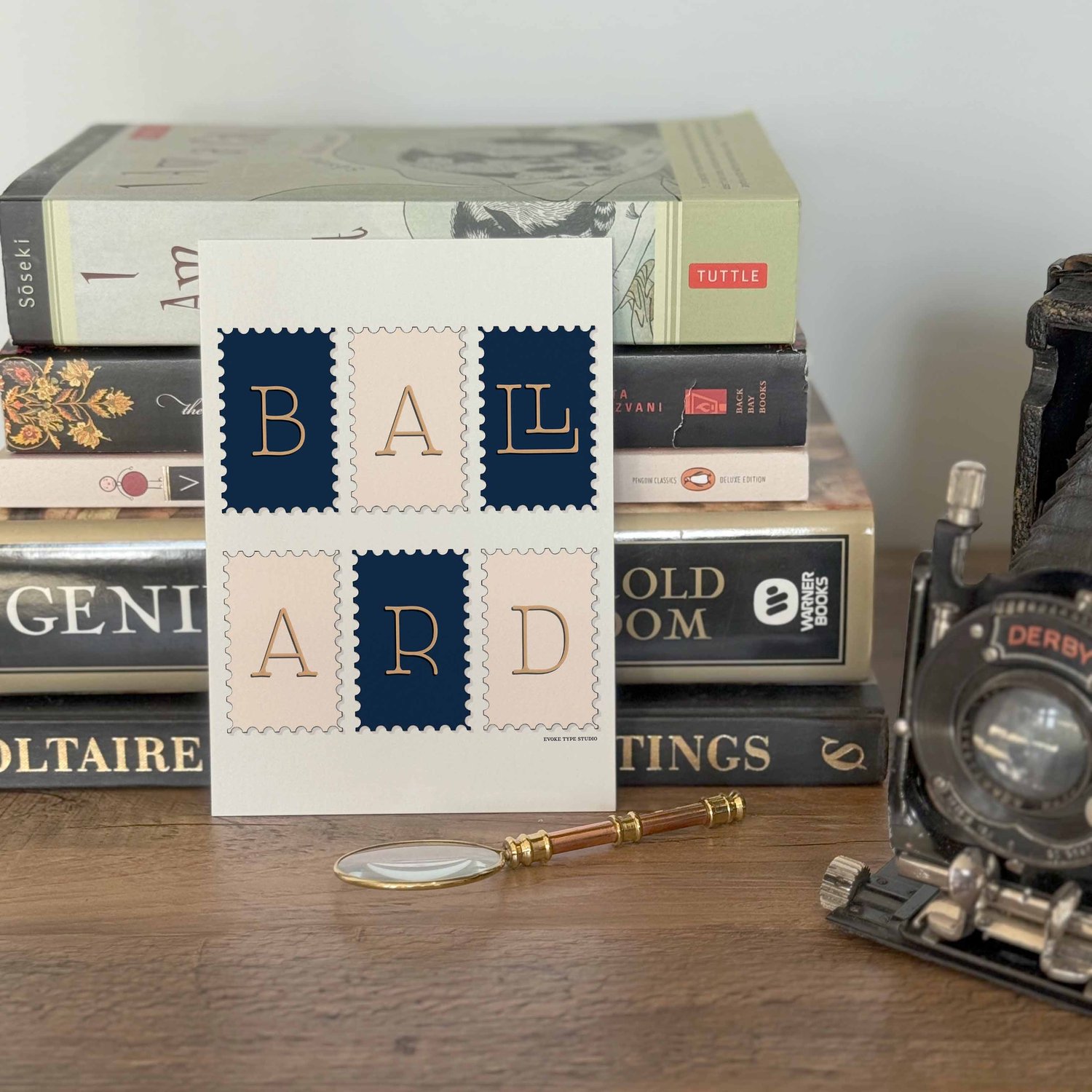 A notecard placed in front of a stack of books with a vintage camera and a small magnifying glass placed nearby. The artwork on the notecard is of postage stamps featuring the Ballard neighborhood in Seattle, Washington drawn in a unique lettering style.
