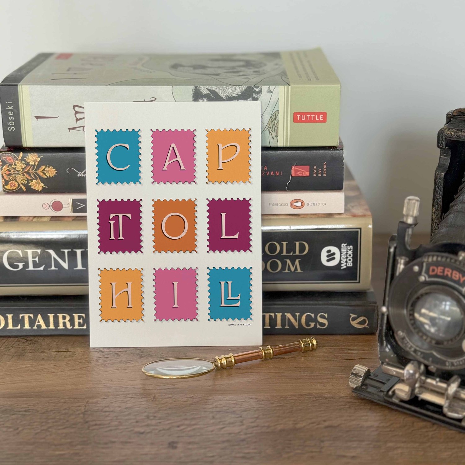 A notecard placed in front of a stack of books with a vintage camera and a small magnifying glass placed nearby. The artwork on the notecard is of postage stamps featuring the Capitol Hill neighborhood in Seattle, Washington drawn in a unique lettering st