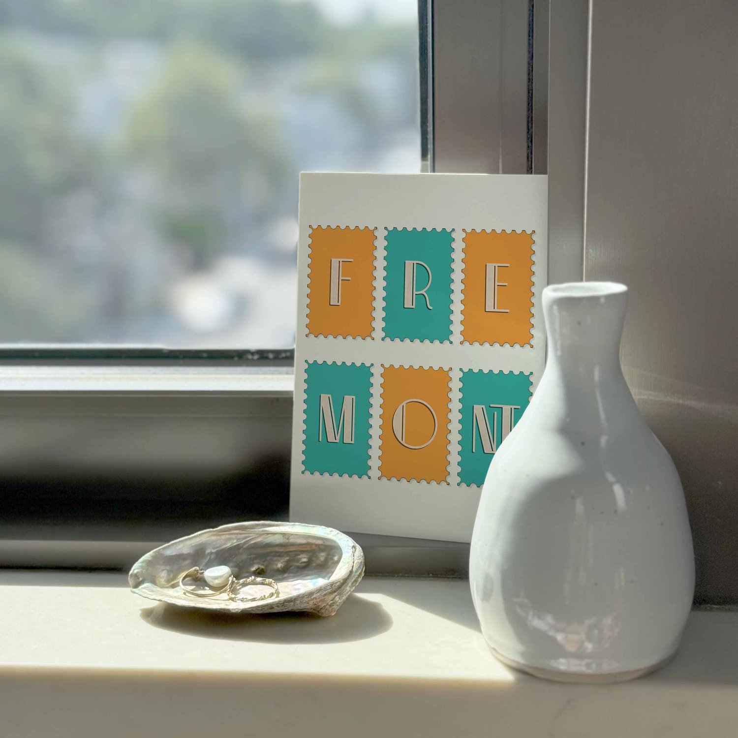 A typography-driven notecard that spells out 'Fremont' on various postage stamps, placed on a windowsill.