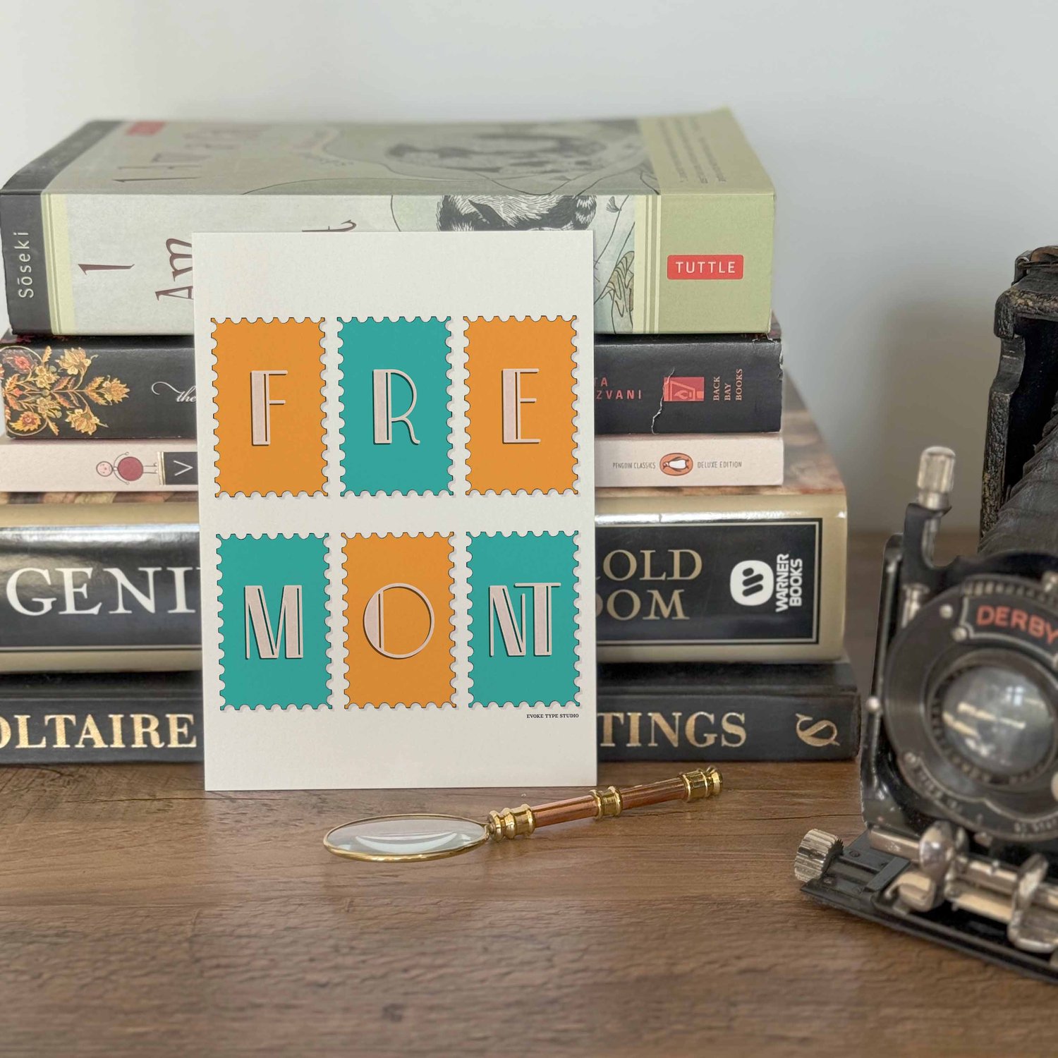 A notecard placed in front of a stack of books with a vintage camera and a small magnifying glass placed nearby. The artwork on the notecard is of postage stamps featuring the Fremont neighborhood in Seattle, Washington drawn in a unique lettering style.