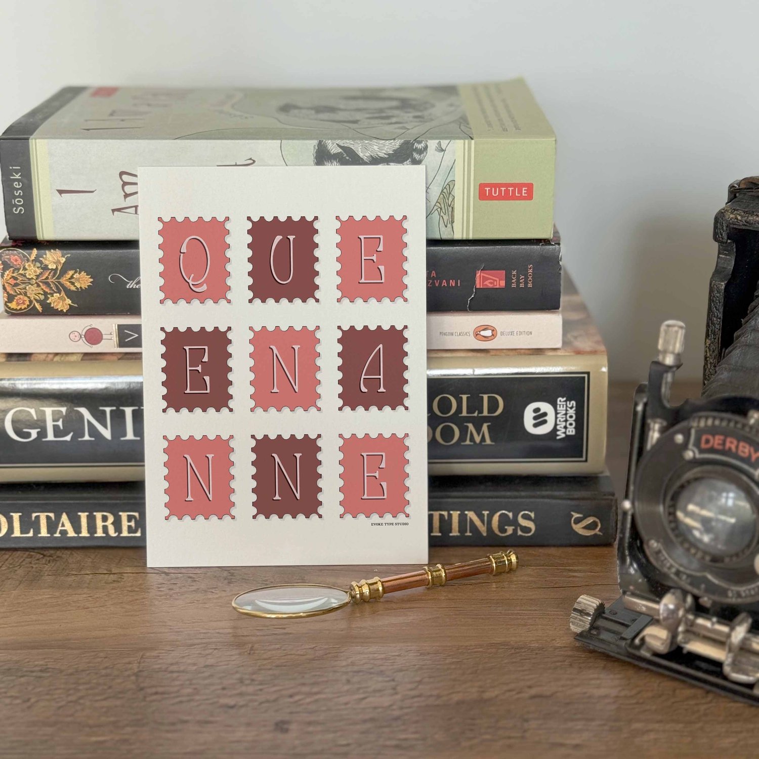 A notecard placed in front of a stack of books with a vintage camera and a small magnifying glass placed nearby. The artwork on the notecard is of postage stamps featuring the Queen Anne neighborhood in Seattle, Washington drawn in a unique lettering styl