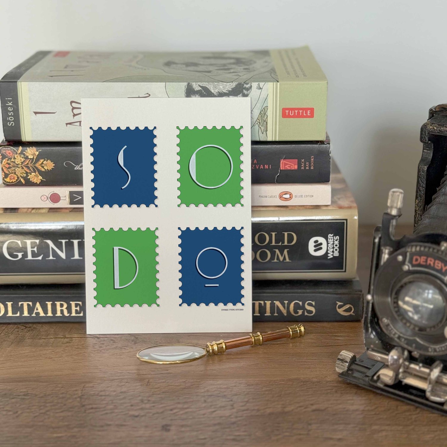 A notecard placed in front of a stack of books with a vintage camera and a small magnifying glass placed nearby. The artwork on the notecard is of postage stamps featuring the SoDo neighborhood in Seattle, Washington drawn in a unique lettering style.