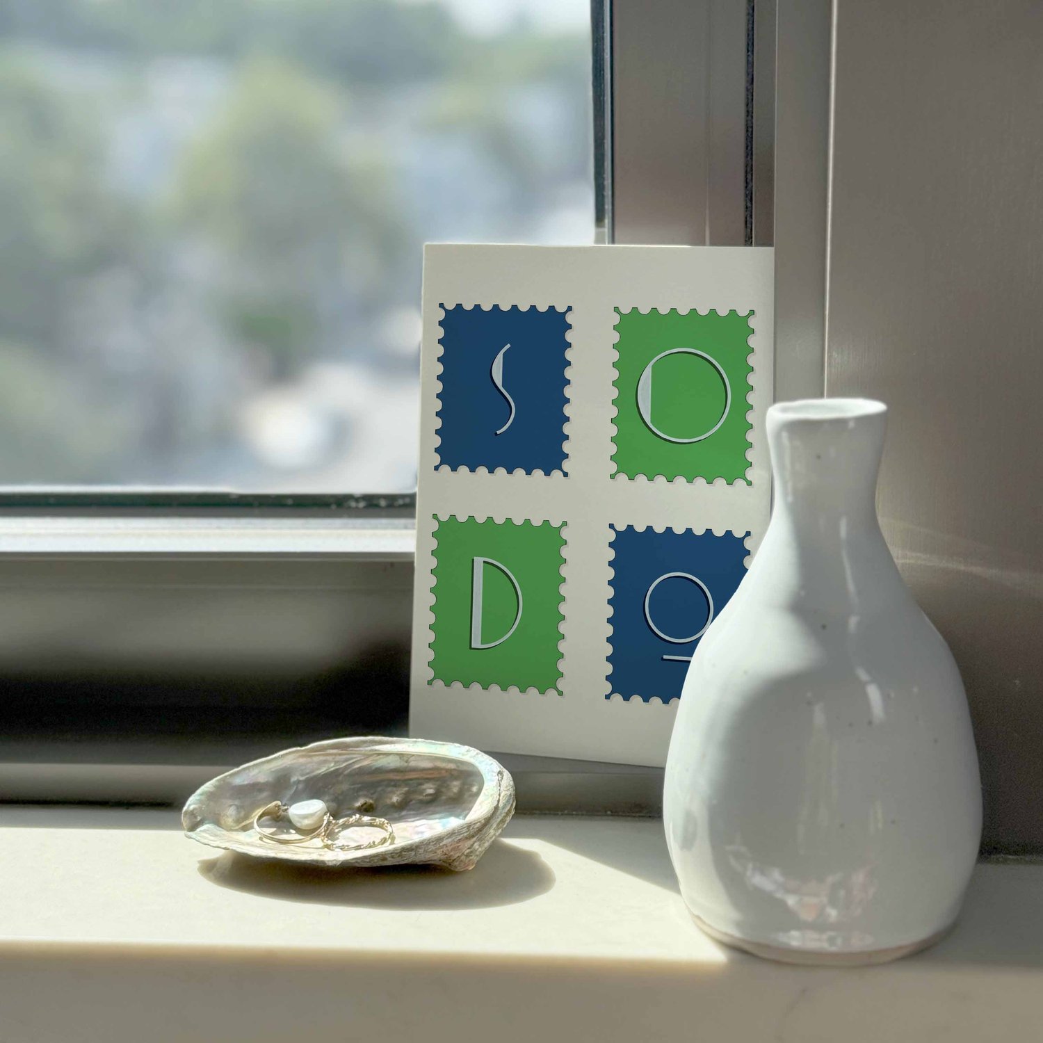 A typography-driven notecard that spells out 'SoDo' on various postage stamps, placed on a windowsill.