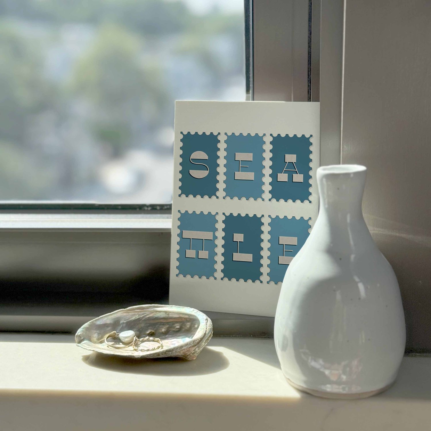 A notecard sitting on a windowsill surrounding by a white vase and a pretty shell with jewelry placed inside.