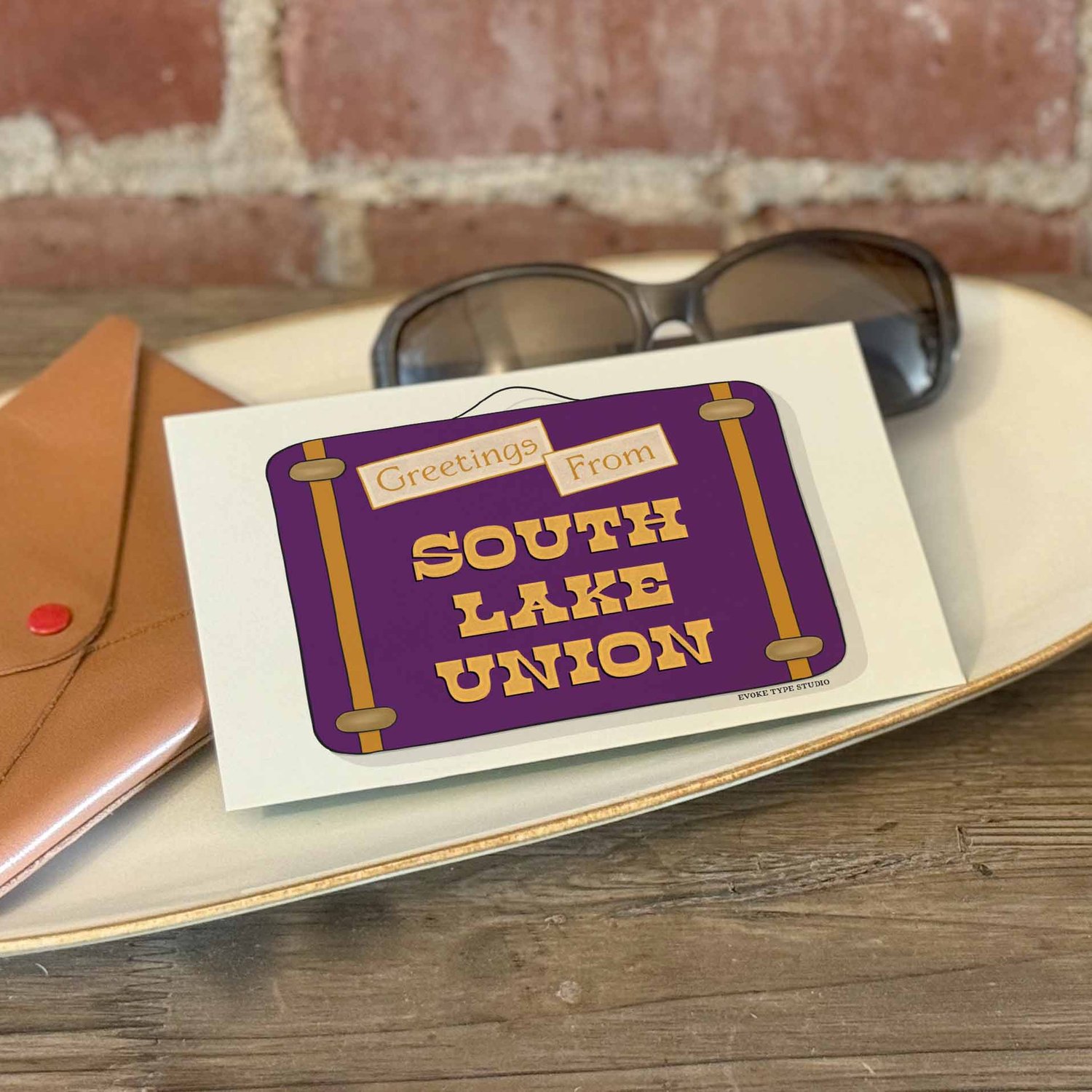 A postcard with a vintage suitcase illustration placed on a bureau with a wallet and sunglasses placed nearby.