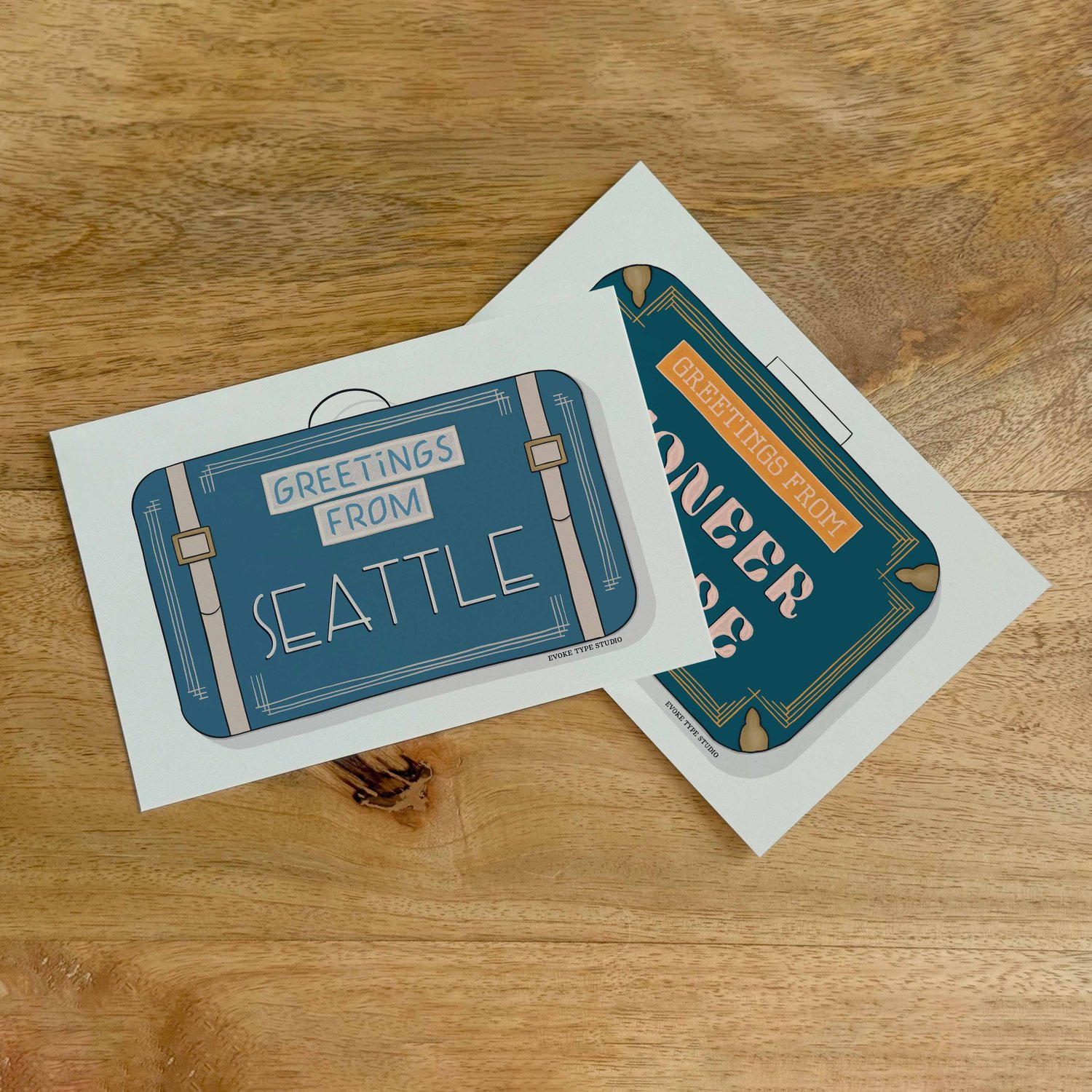Two postcards, one placed on top of the other. Both postcards feature vintage suitcase illustrations and are for the Seattle, Washington area.