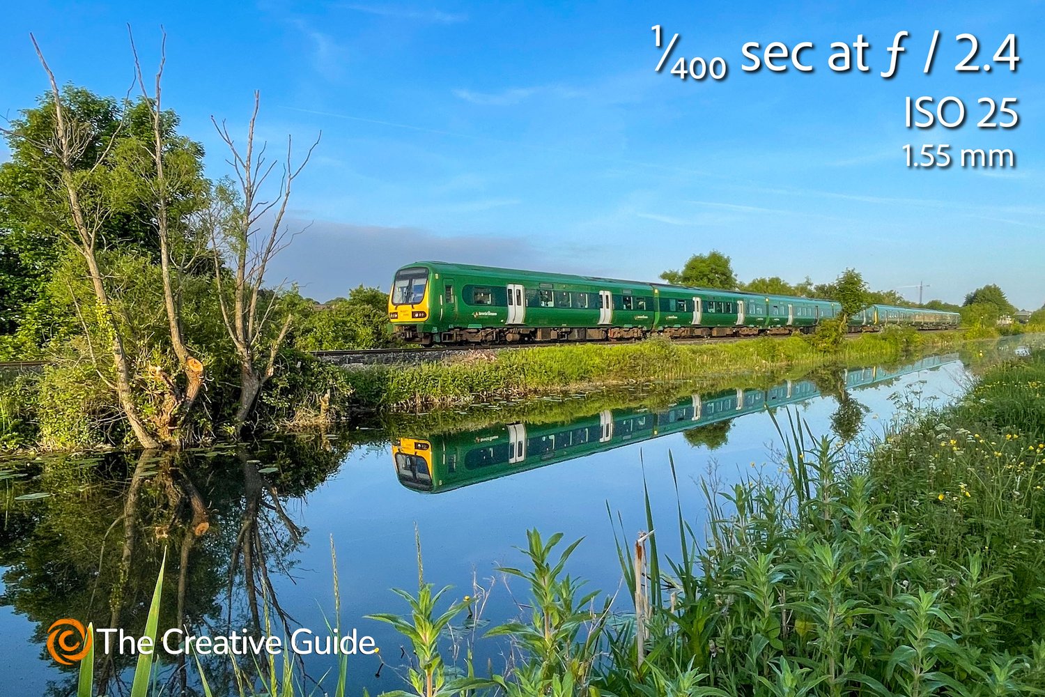 Green commuter train traveling along tracks with a perfect reflection in calm water, surrounded by summer greenery, photographed at 1/400 sec f/2.4 ISO 25, 1.55 mm, The Creative Guide Photo Gallery.