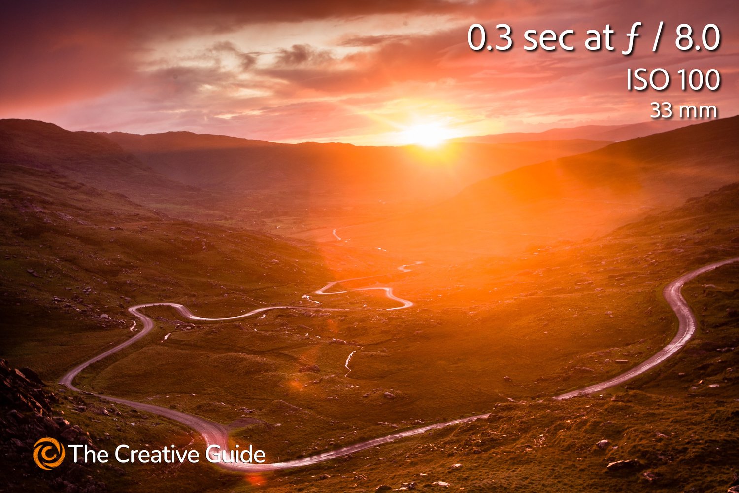 Glowing sunset landscape with winding road through green valleys and hills, bathed in orange light, photographed at 0.3 sec f/8 ISO 100, 33 mm, The Creative Guide Photo Gallery.