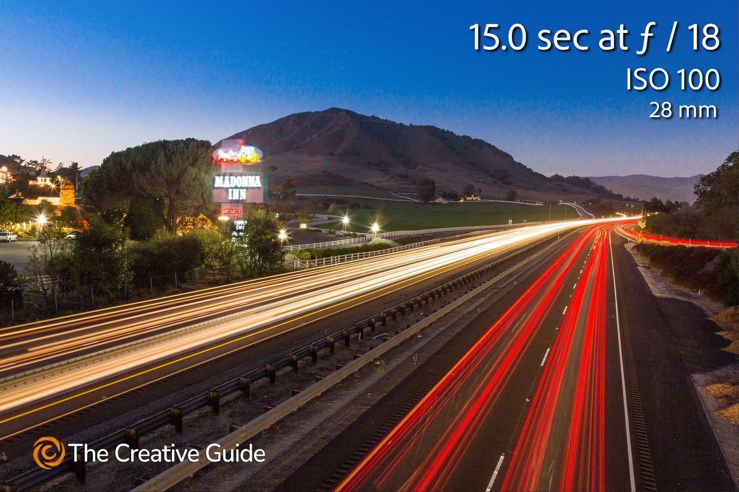 Long exposure photo of a highway at night with red and white light trails from cars, Madonna Inn sign and mountain in background, photographed at 15 sec f/18 ISO 100, 28 mm, The Creative Guide Photo Gallery.