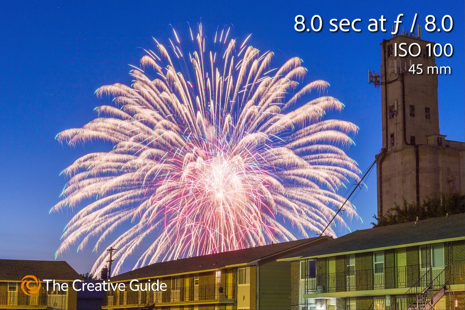 Long exposure fireworks display lighting up the night sky above apartments and a tall concrete tower, photographed at 8 sec f/8 ISO 100, 45 mm, The Creative Guide Photo Gallery.