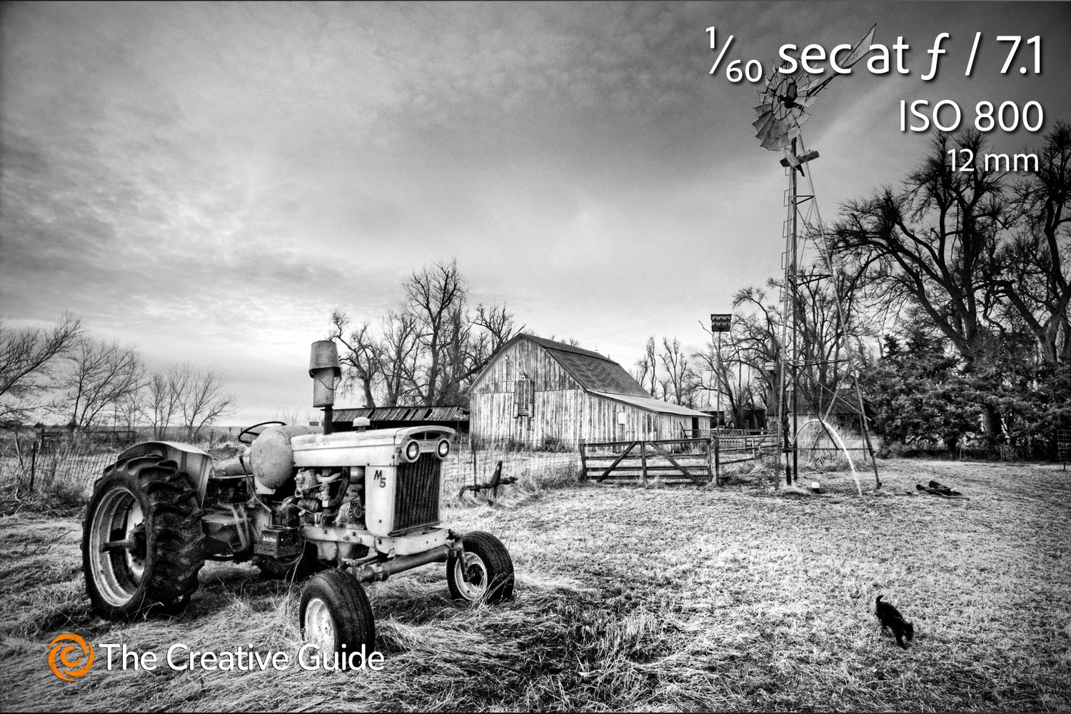 Black and white rural scene with old tractor, weathered barn, and windmill on farmland, photographed at 1/60 sec f/7.1 ISO 800, 12 mm, The Creative Guide Photo Gallery.